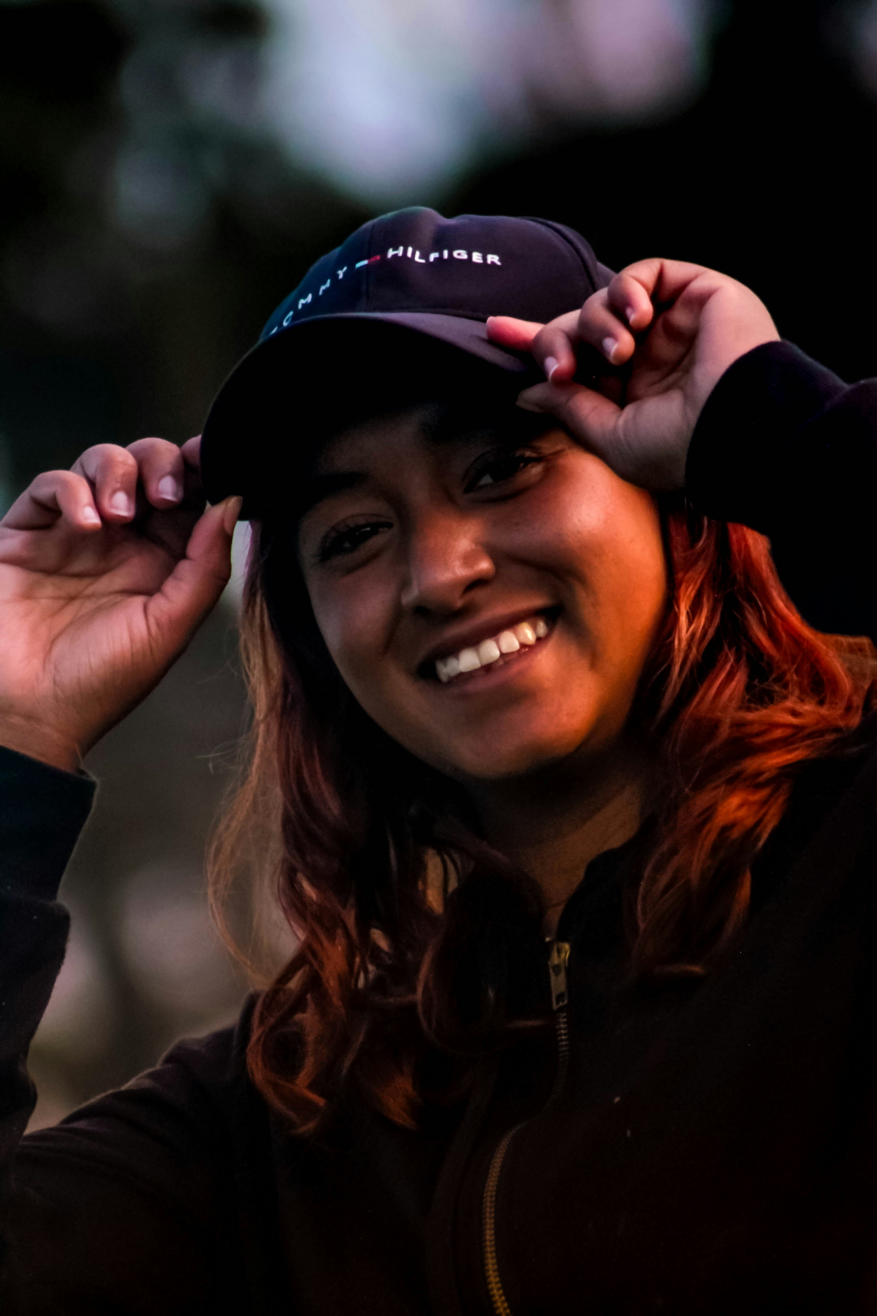 A smiling person with reddish hair wearing a dark baseball cap