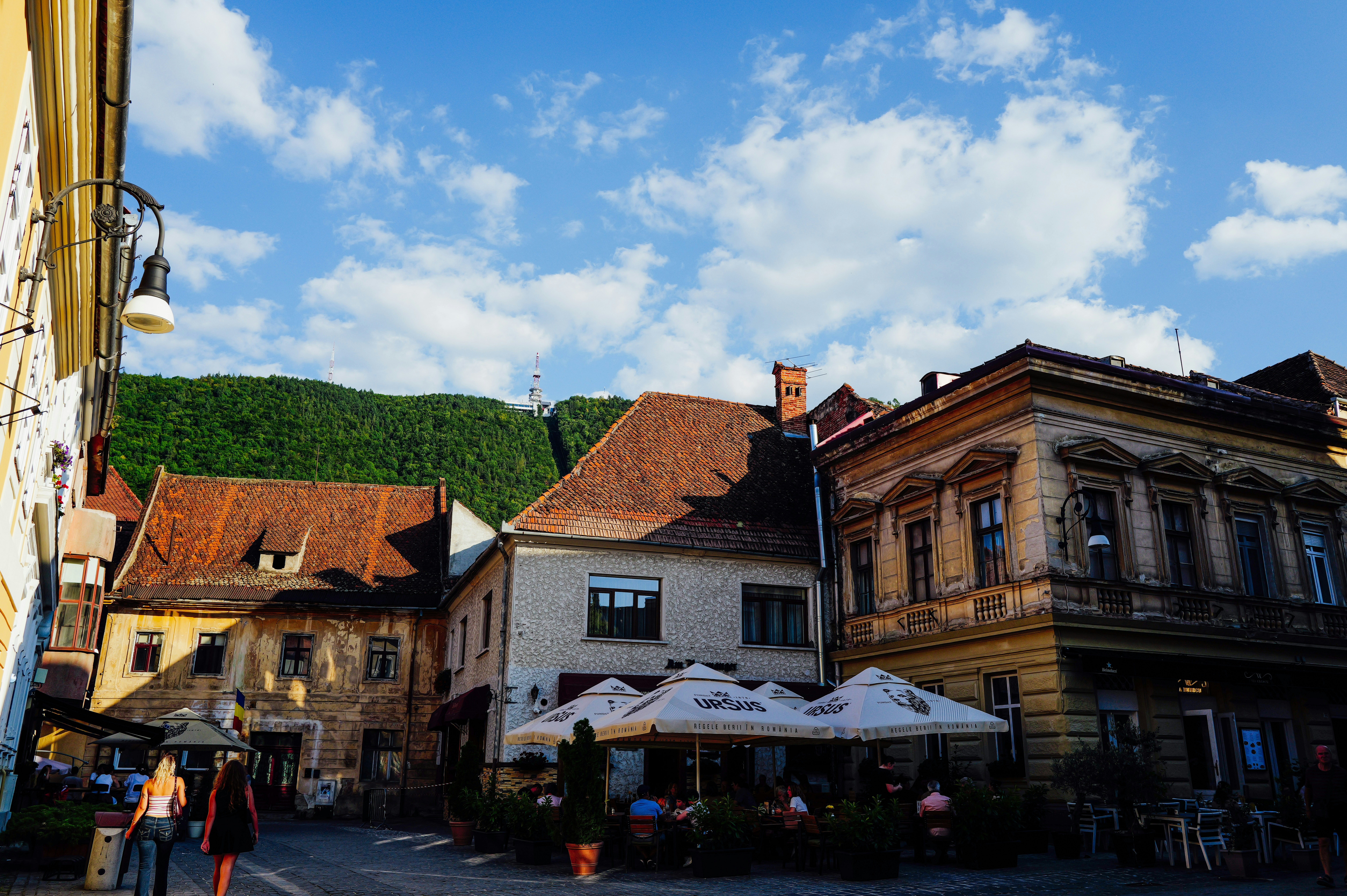 People at outdoor cafe tables with umbrellas beneath a forested mountain in Brasov