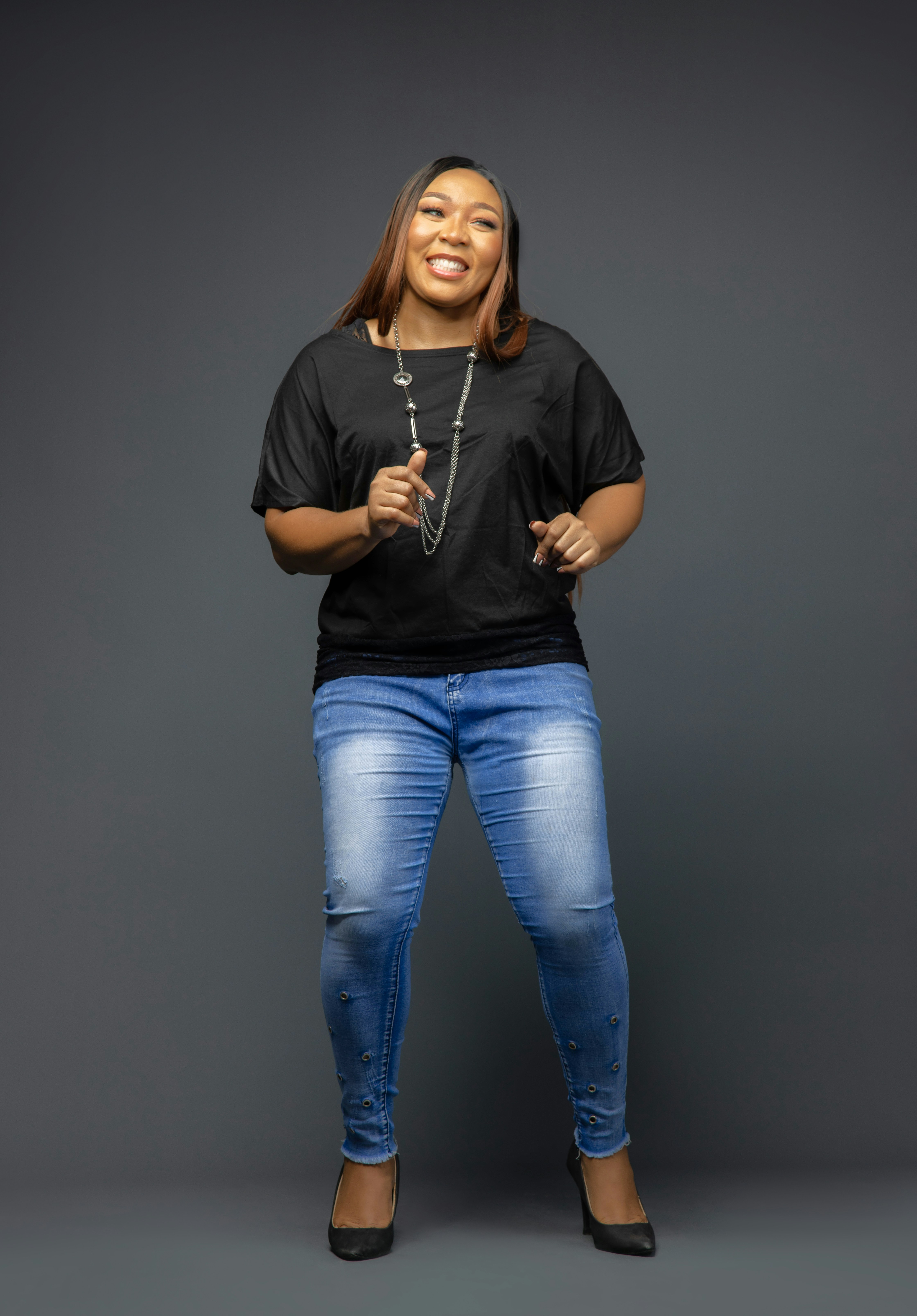 A smiling woman in a black top and blue jeans against a grey background