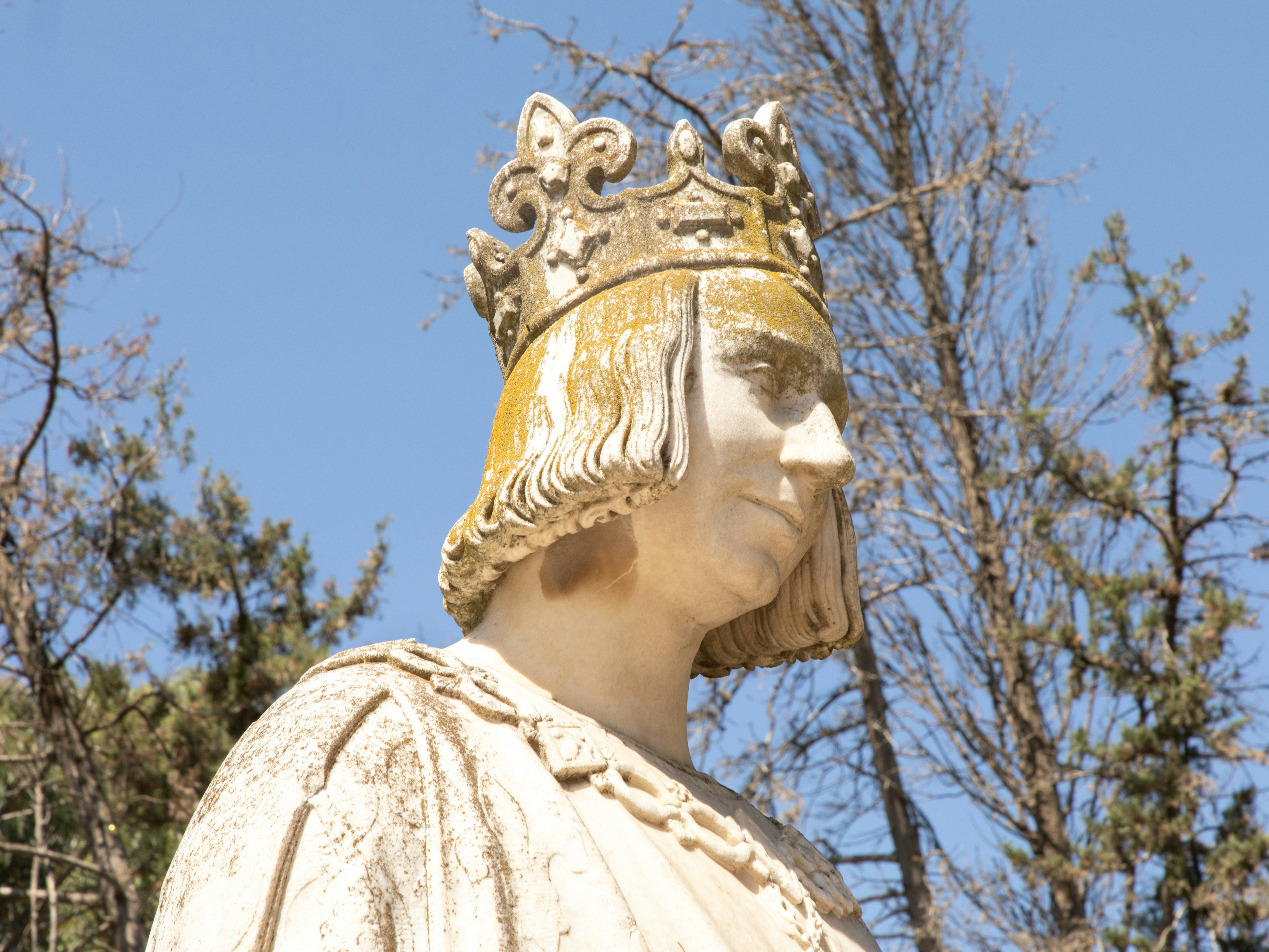 A weathered stone statue of a king wearing a crown against bare trees