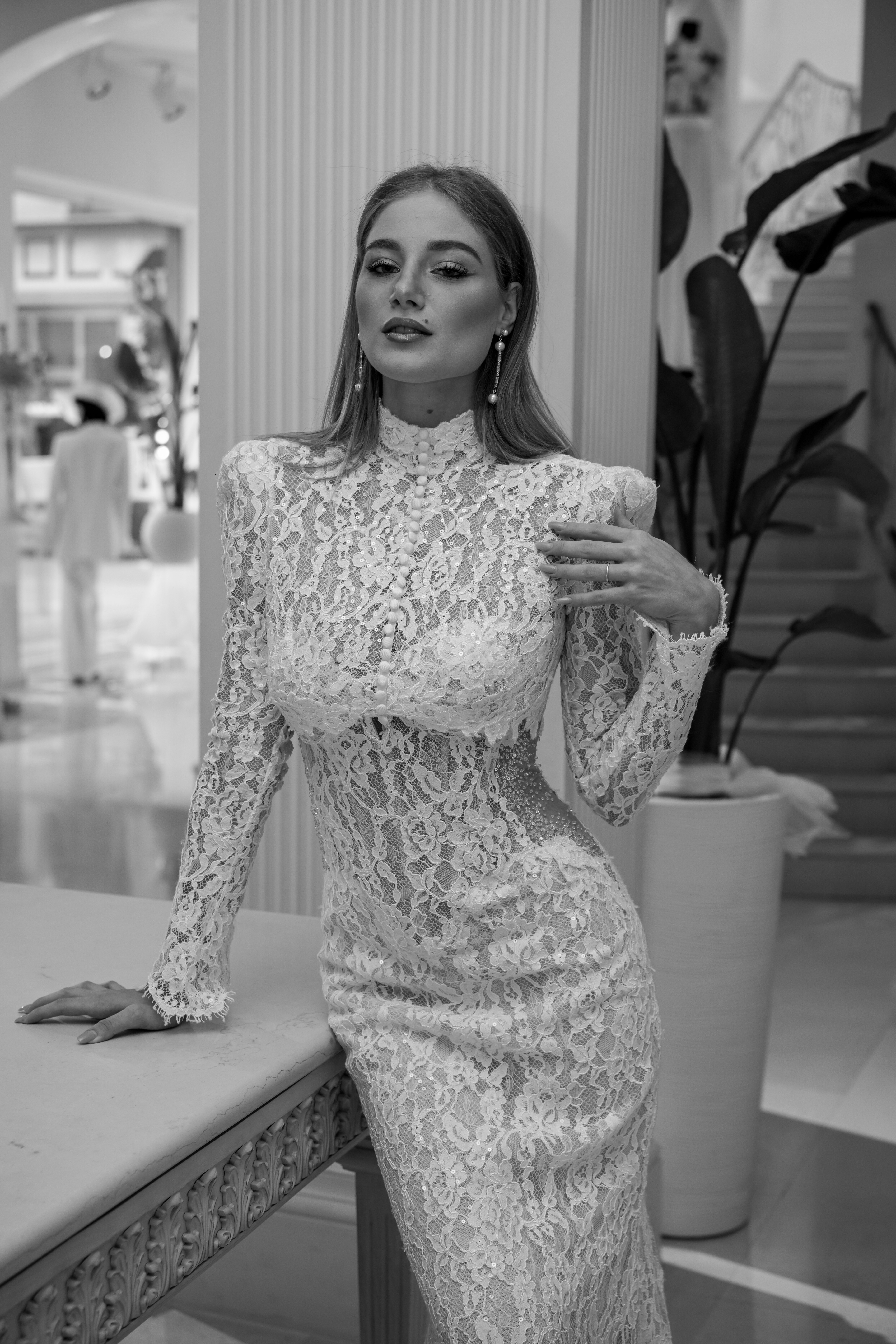 A woman in a white lace dress leaning against a table in a room