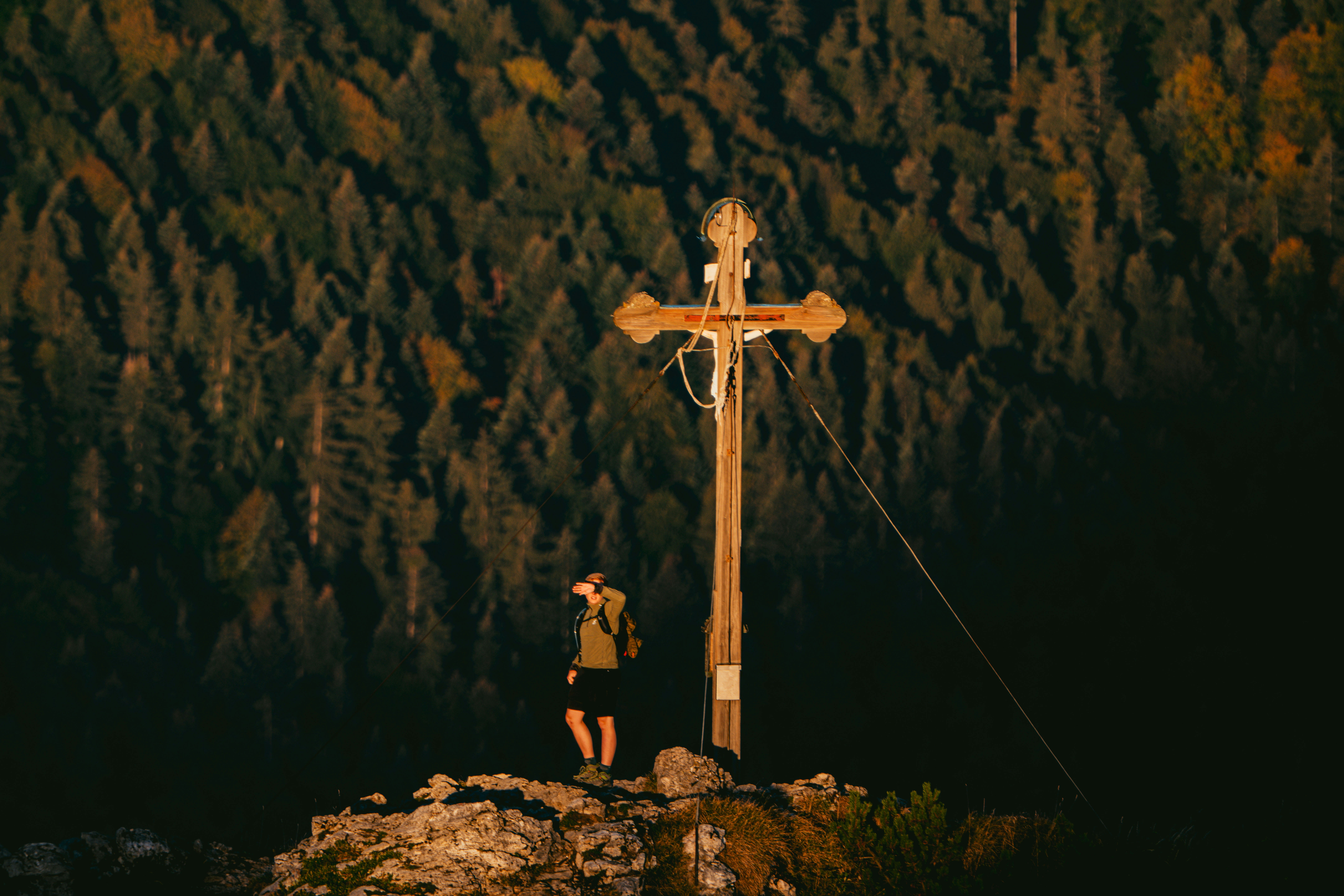 A person shielding eyes from low sun beside a wooden cross on a mountain