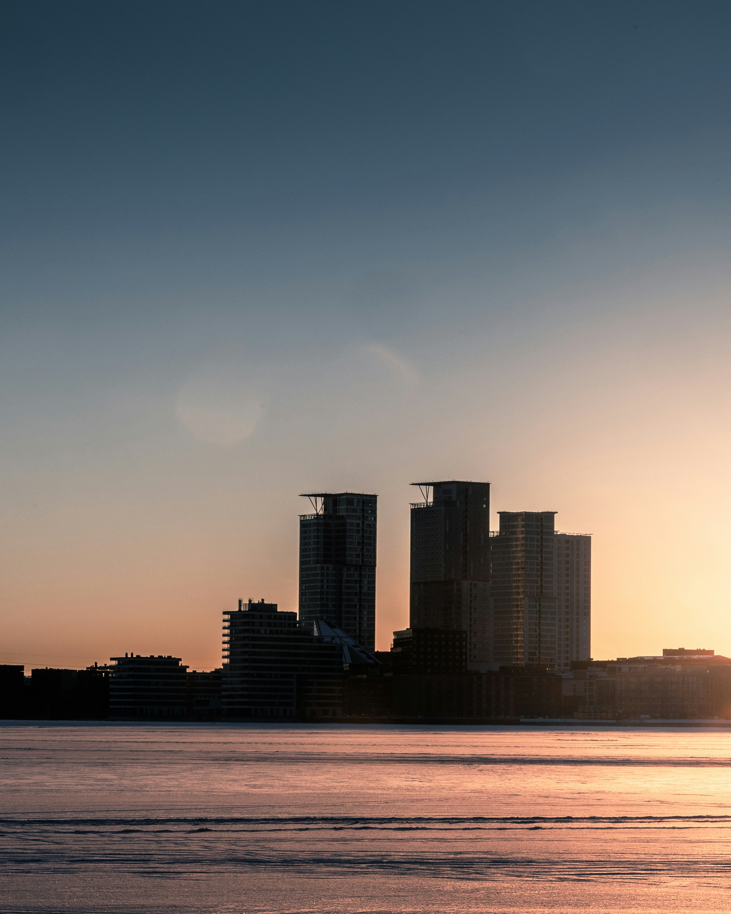 Silhouetted skyscrapers against a warm orange sky above a frozen harbor