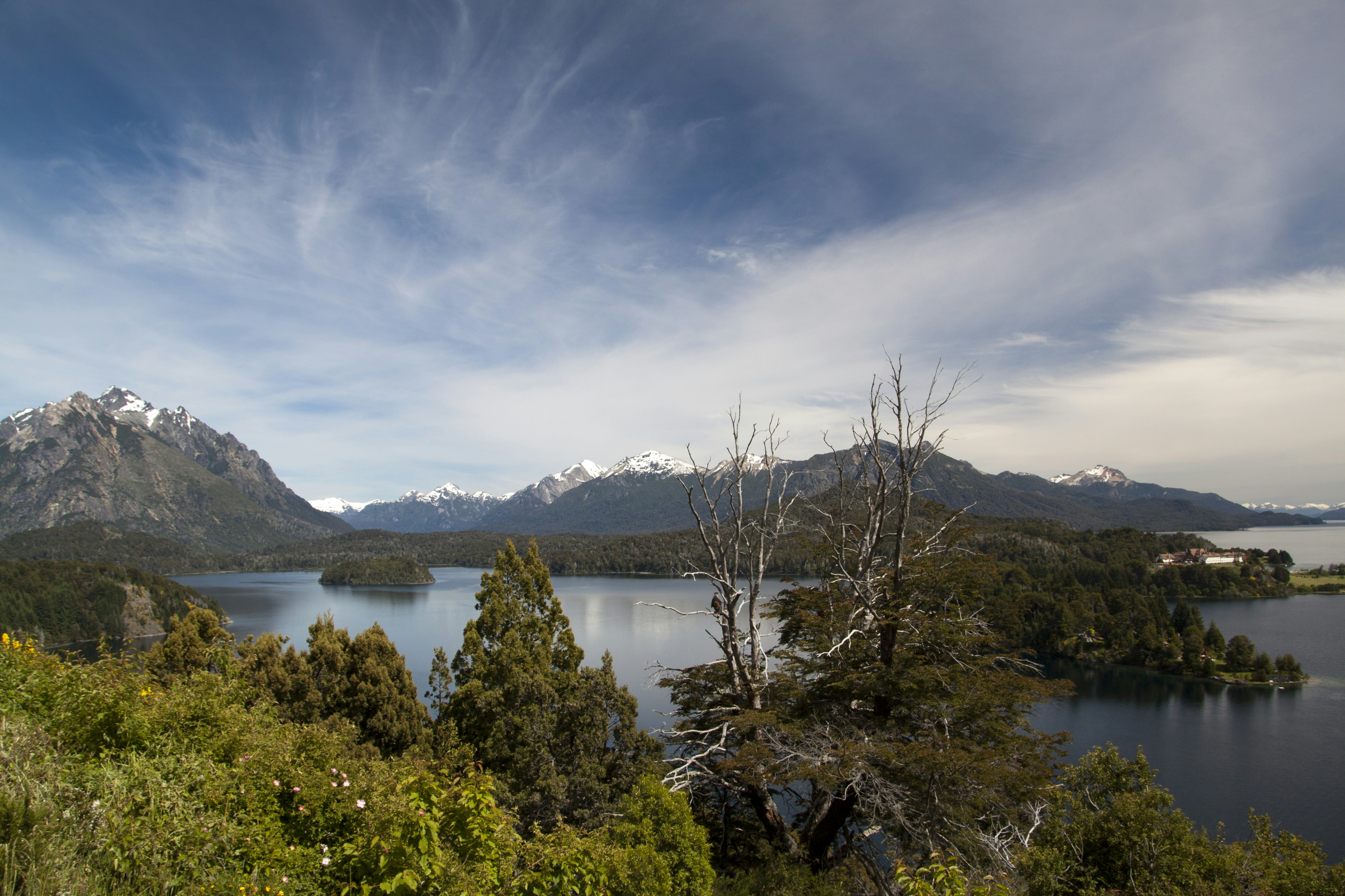 A calm lake surrounded by forested hills and snow-capped mountains under a cloudy sky