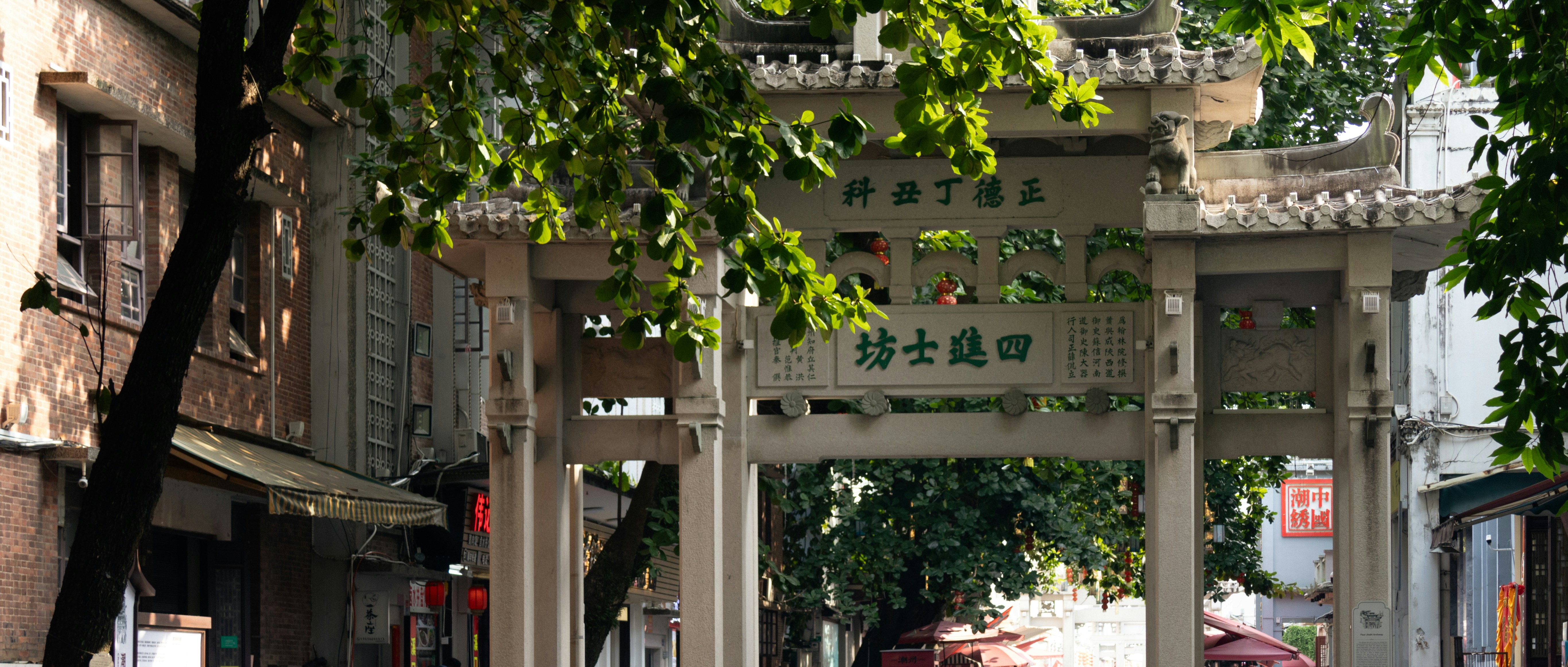 A stone paifang gate with Chinese characters on a street shaded by trees