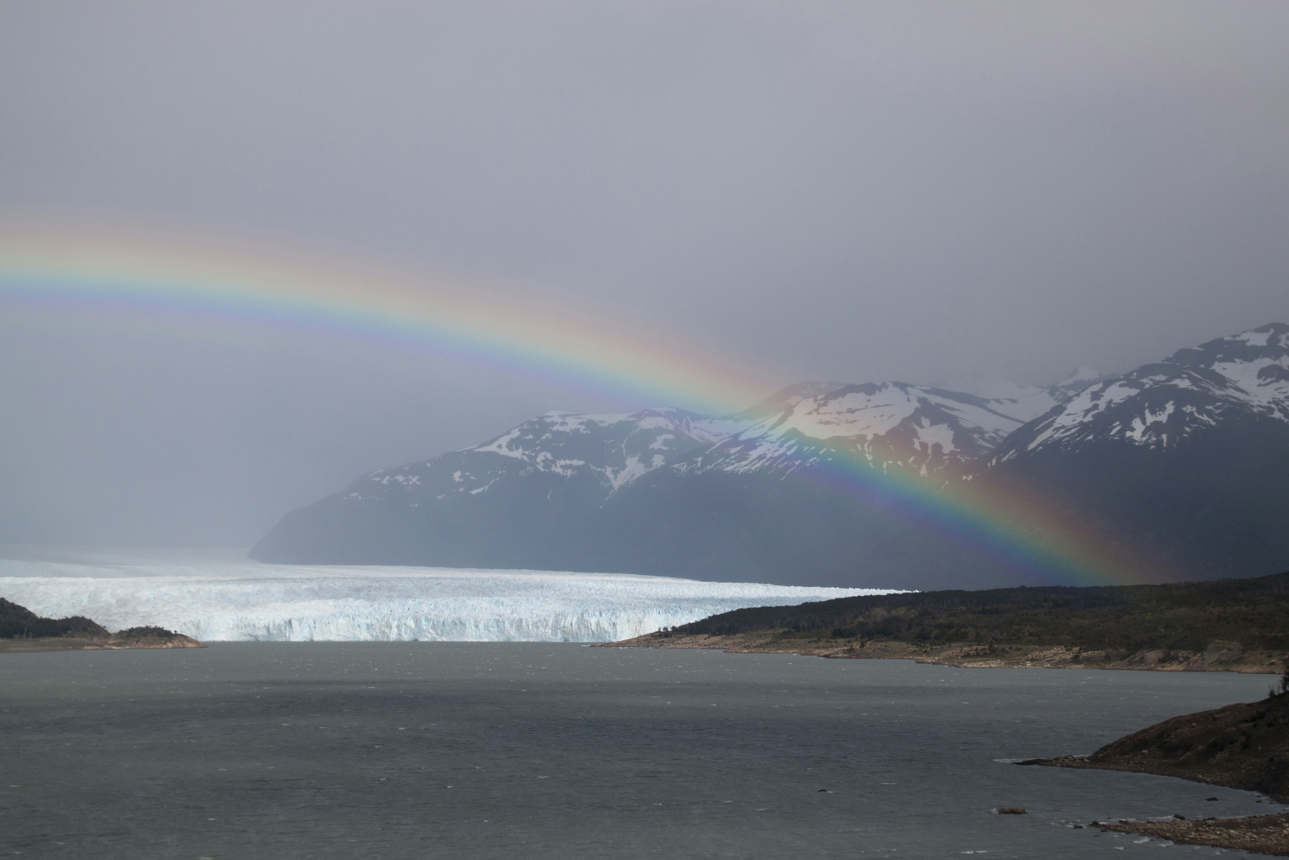 A rainbow arching over a glacier and snow-capped mountains above a lake