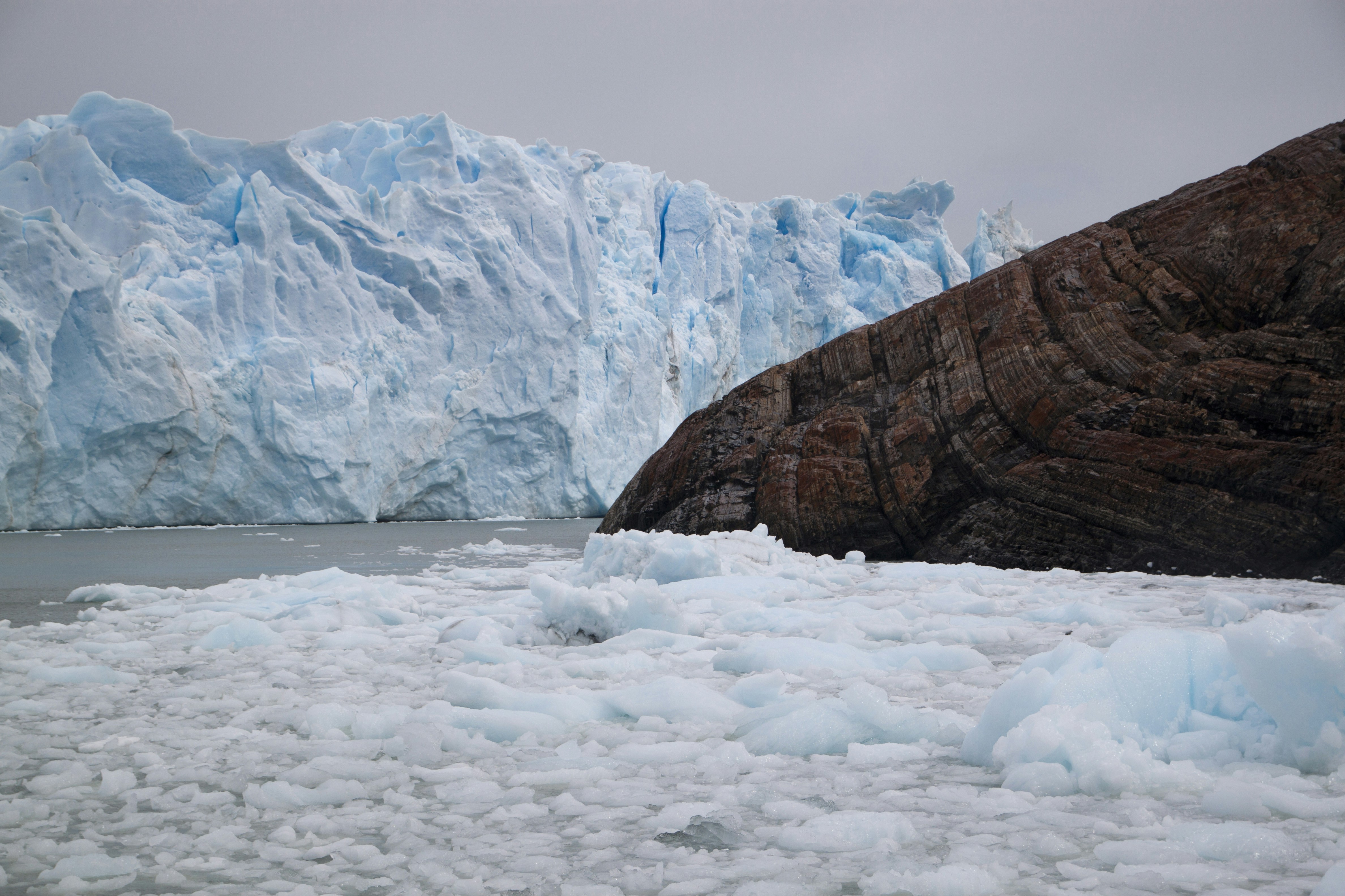 A massive blue glacier wall beside a dark rocky cliff with floating ice chunks