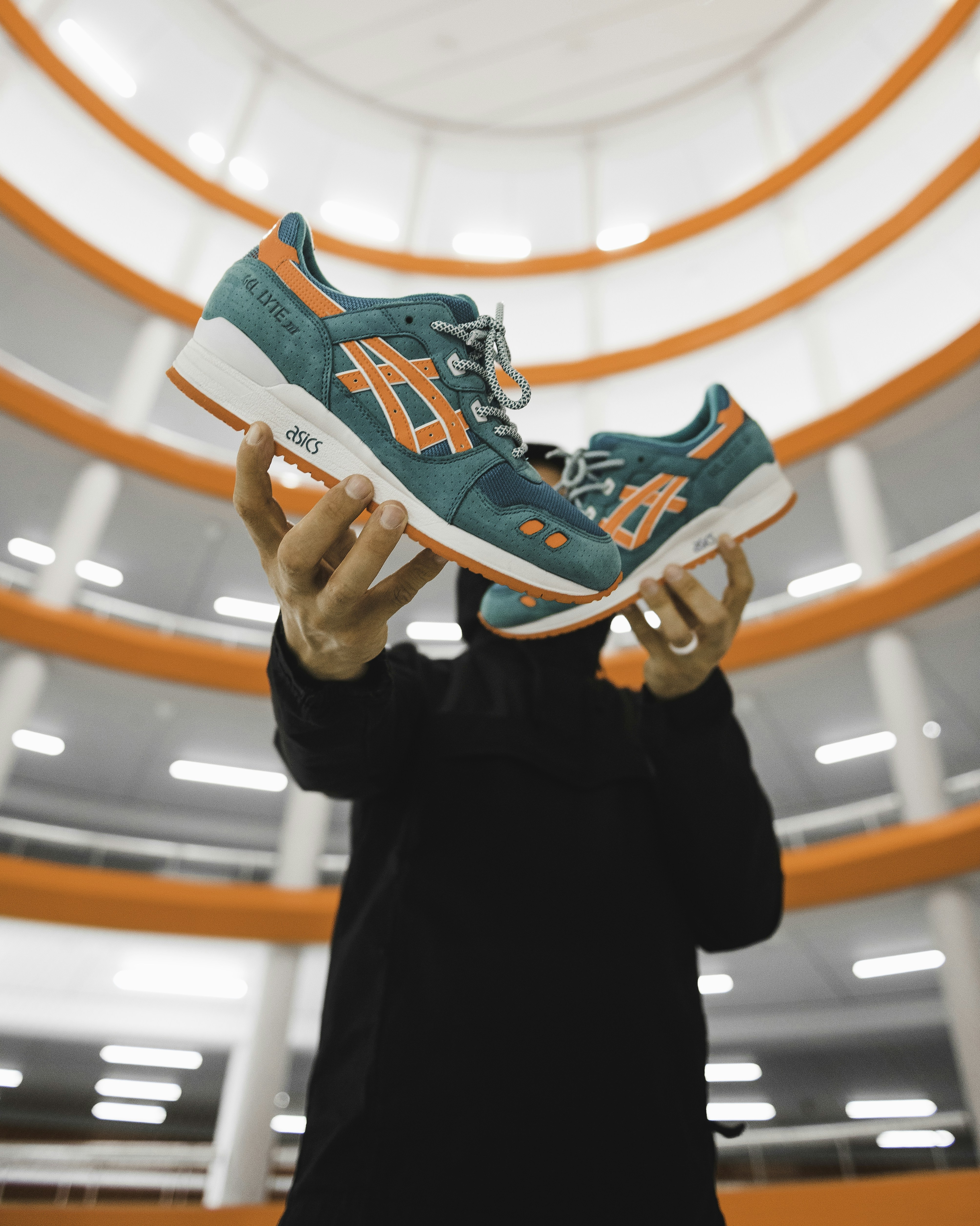A person in a black hoodie holding two teal and orange sneakers