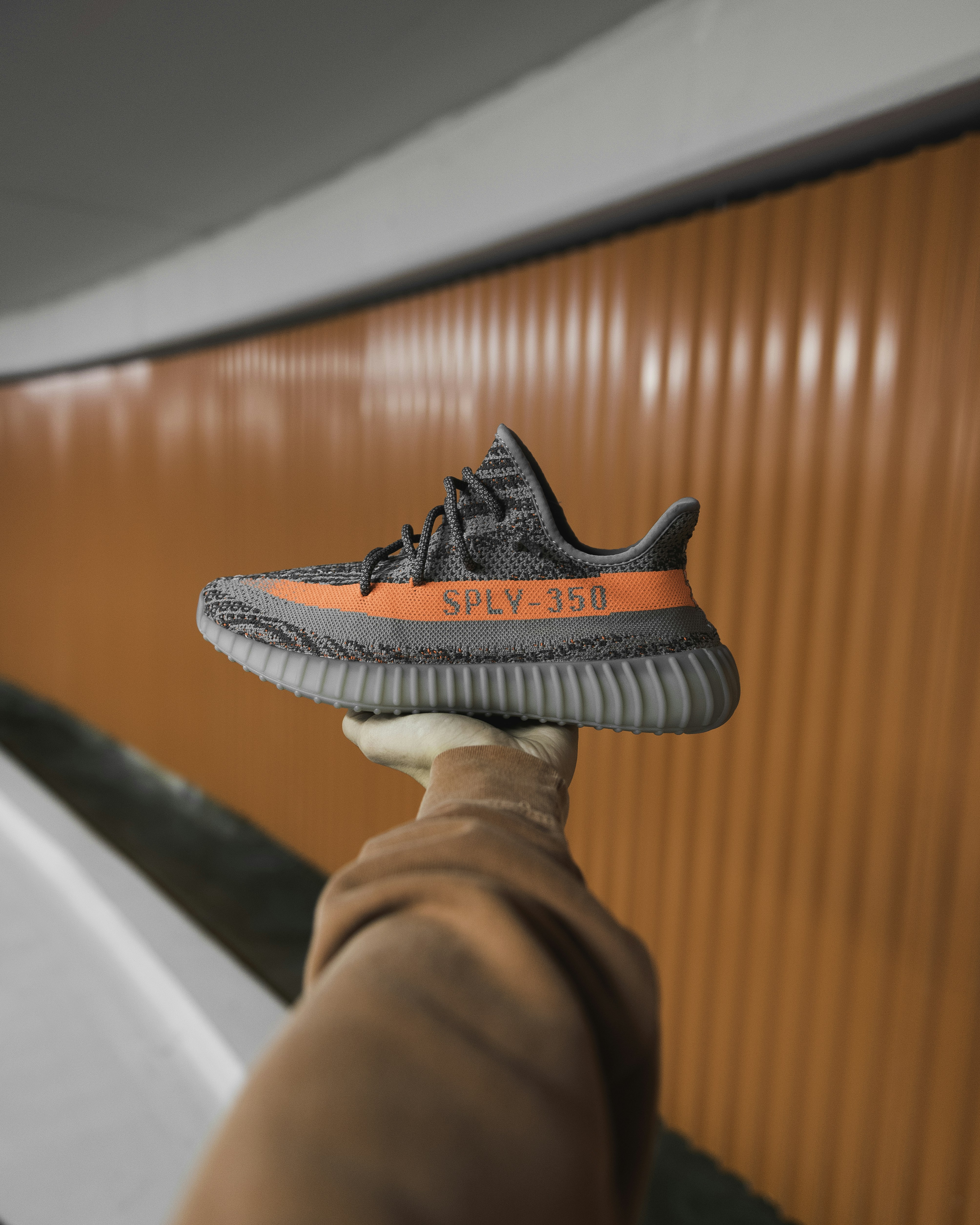 A hand holding a grey sneaker with a bright orange stripe against an orange wall