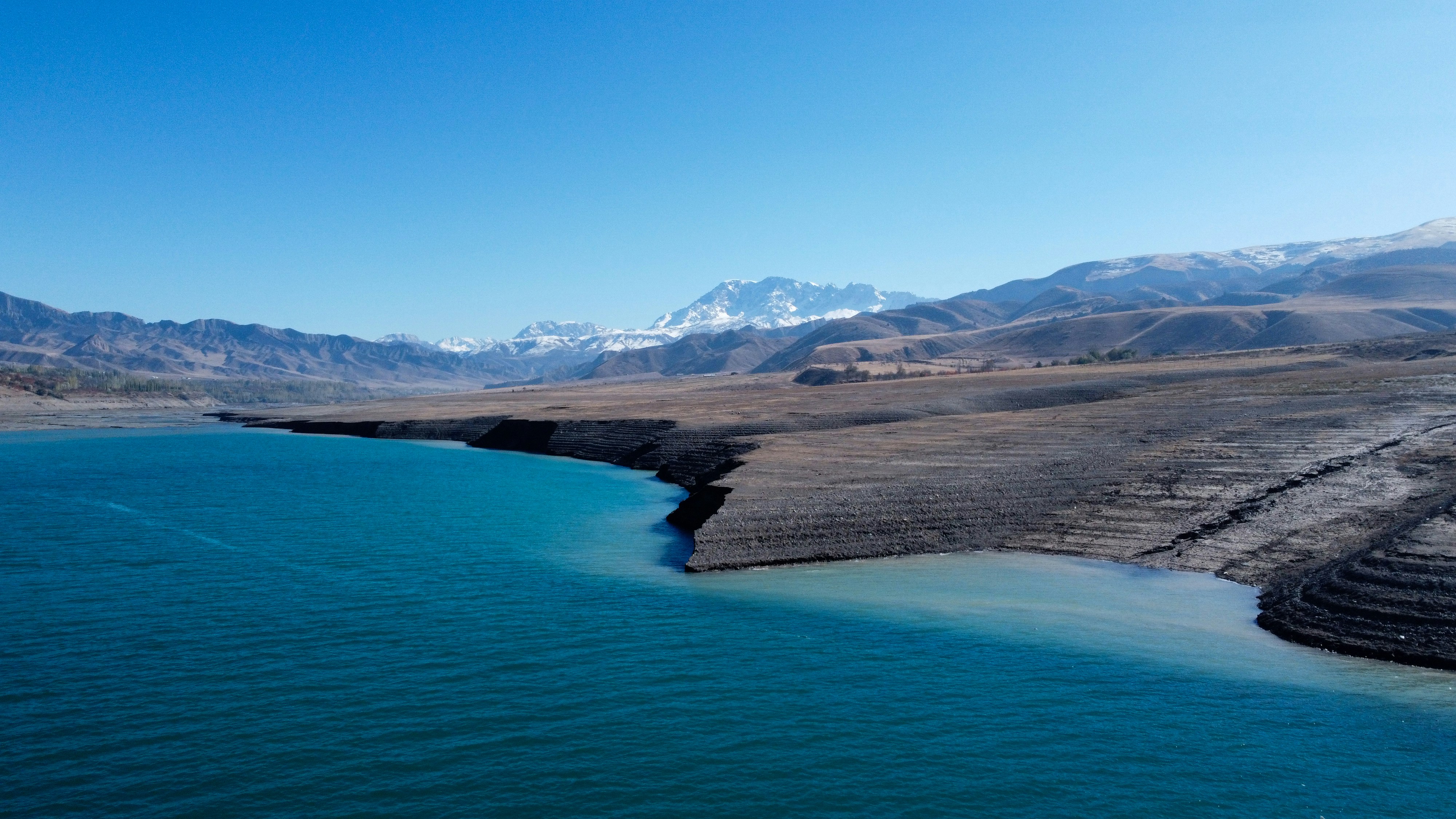 A turquoise lake beside a dry brown shoreline with snow-capped mountains in the distance