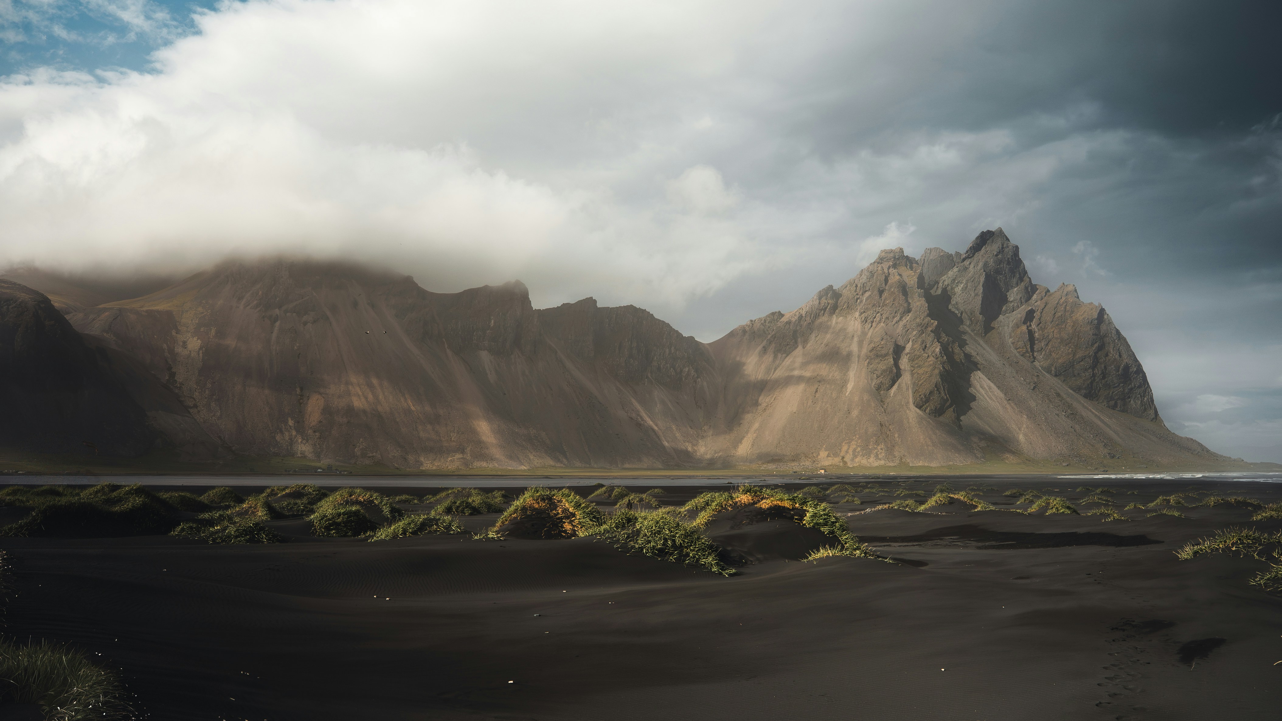 A black sand beach with green grass mounds below jagged mountains under clouds