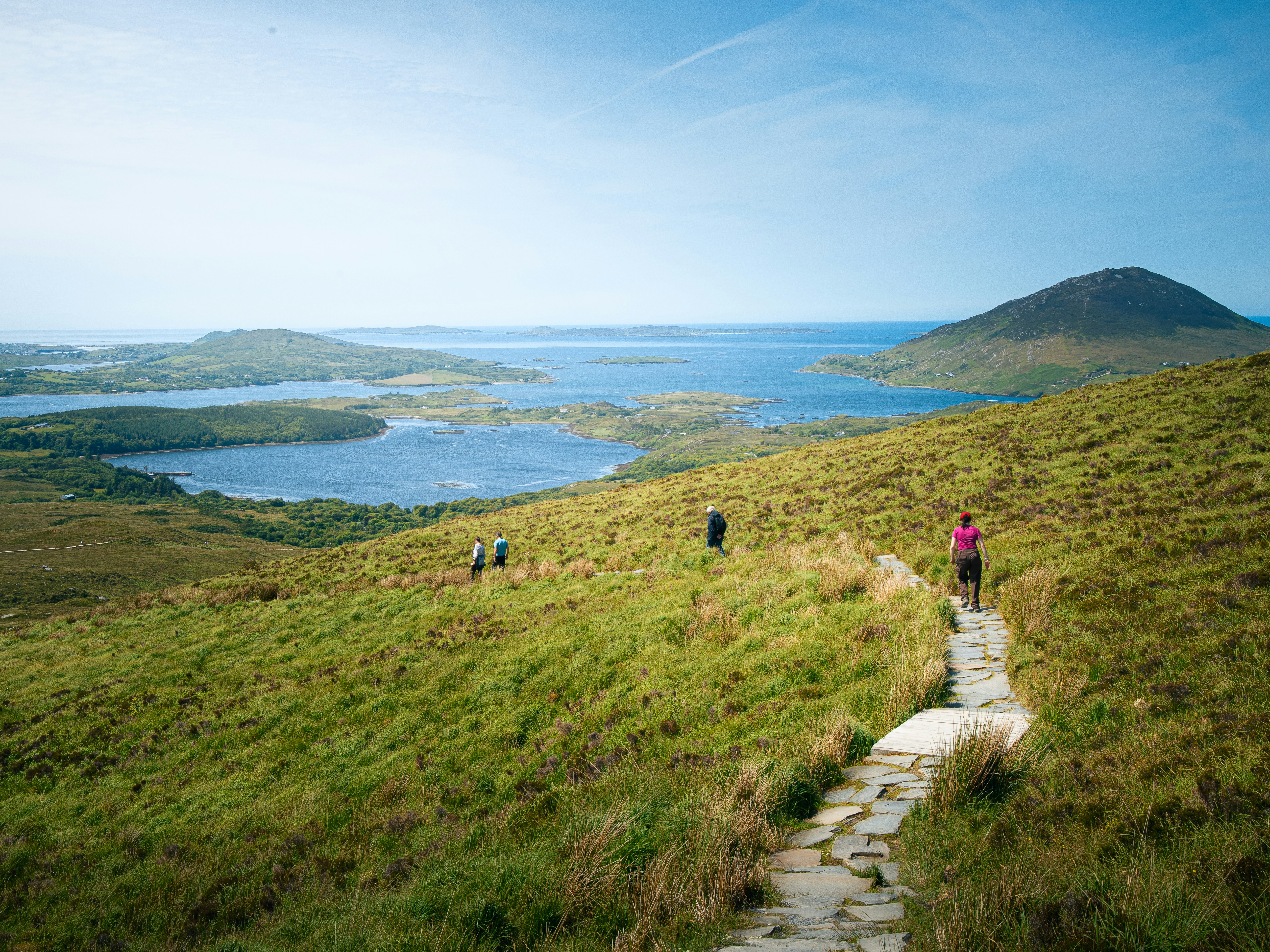 People walking along a stone path through a grassy hillside overlooking a bay