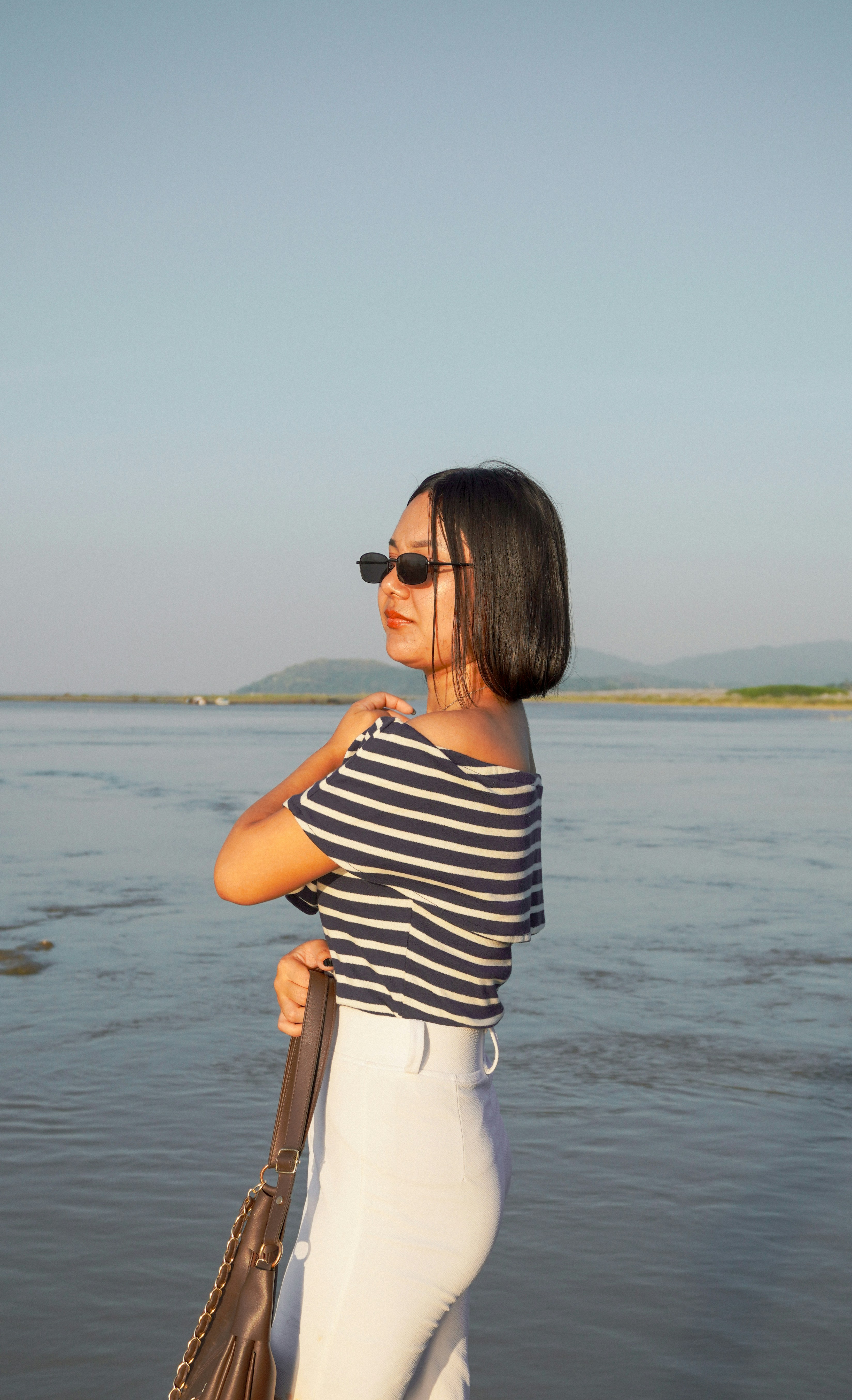 A woman in a striped off-shoulder top and white pants by a river