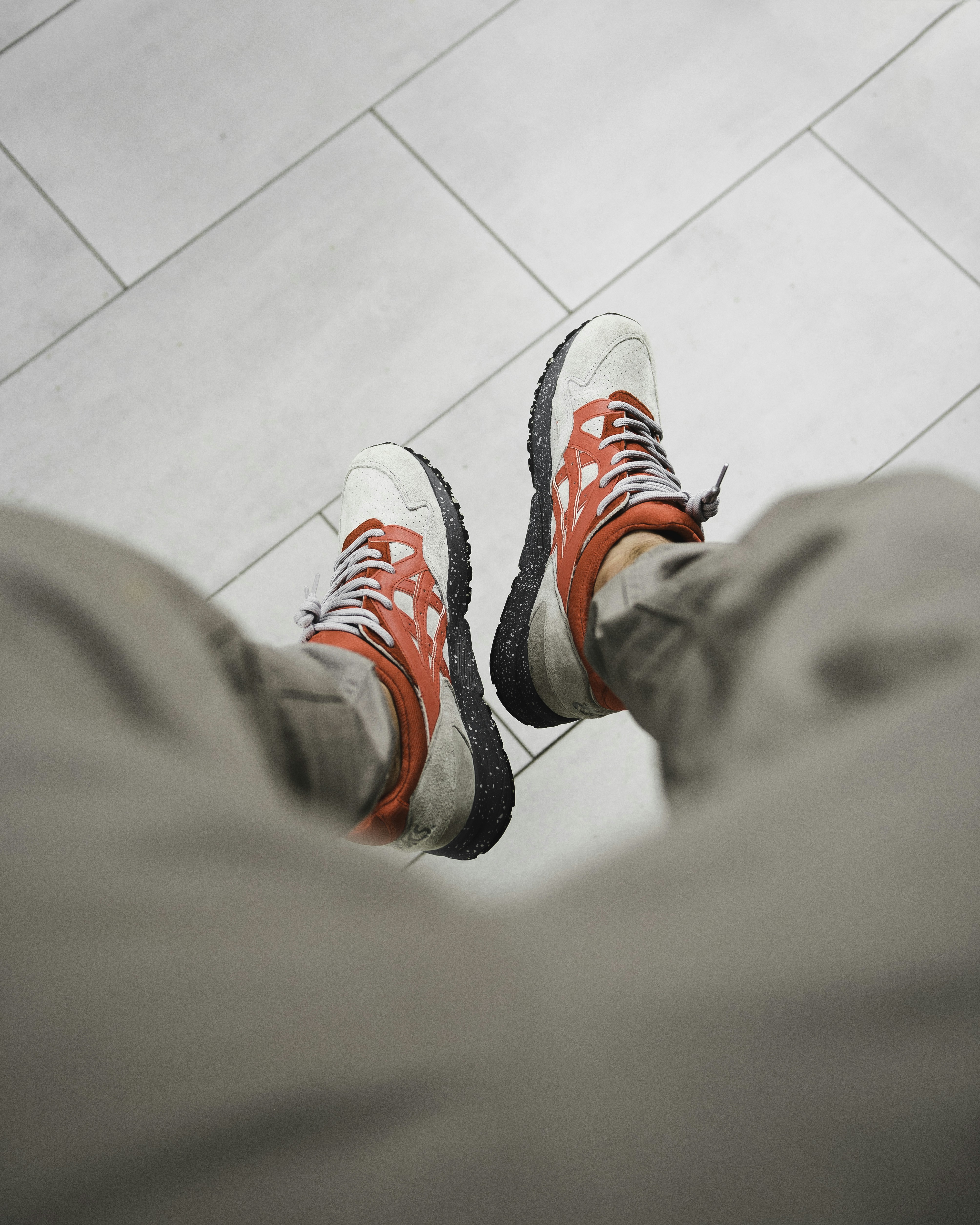 A person wearing red and grey sneakers with speckled soles on a tiled floor