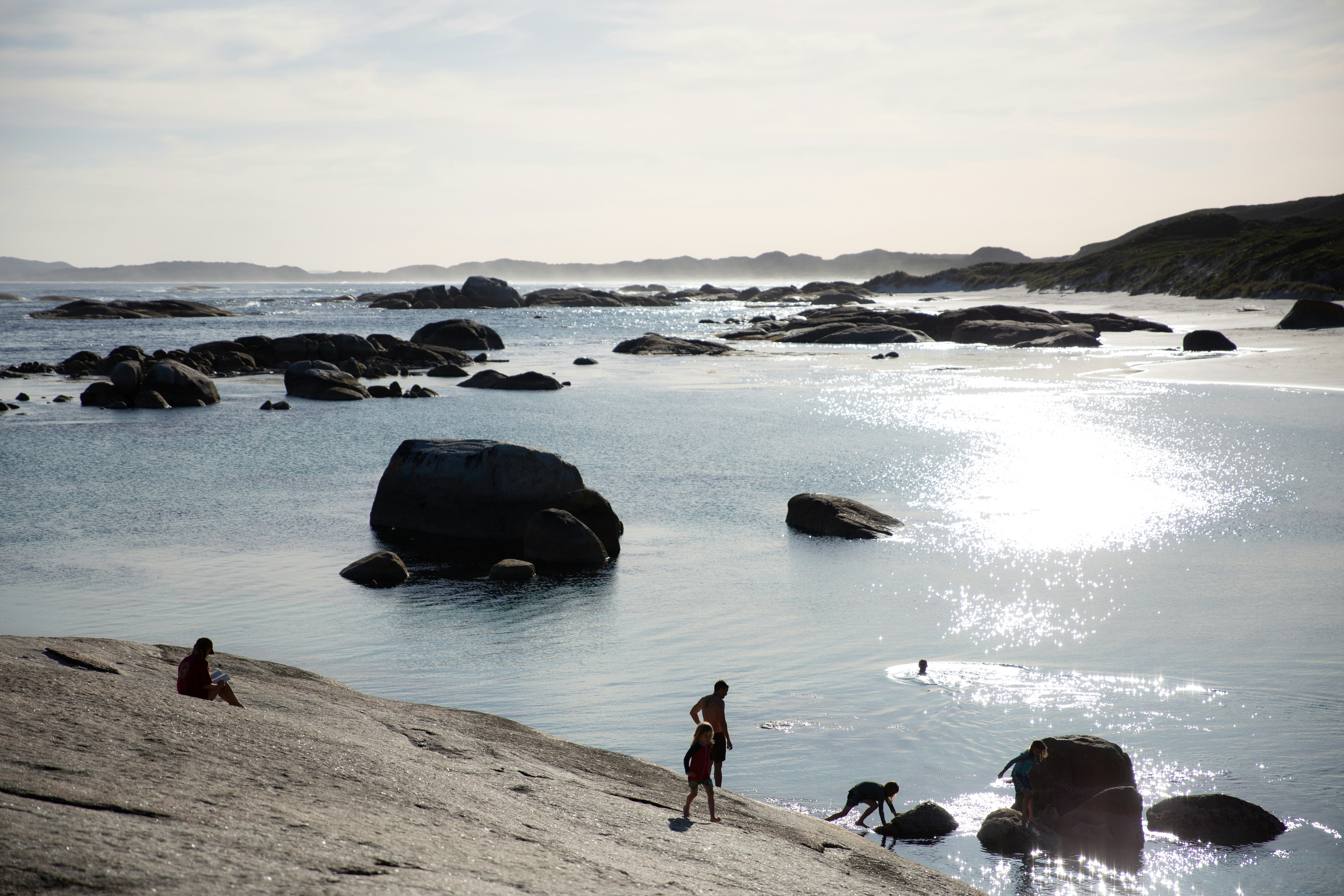 People on a large granite rock by a sunlit coastal bay with boulders