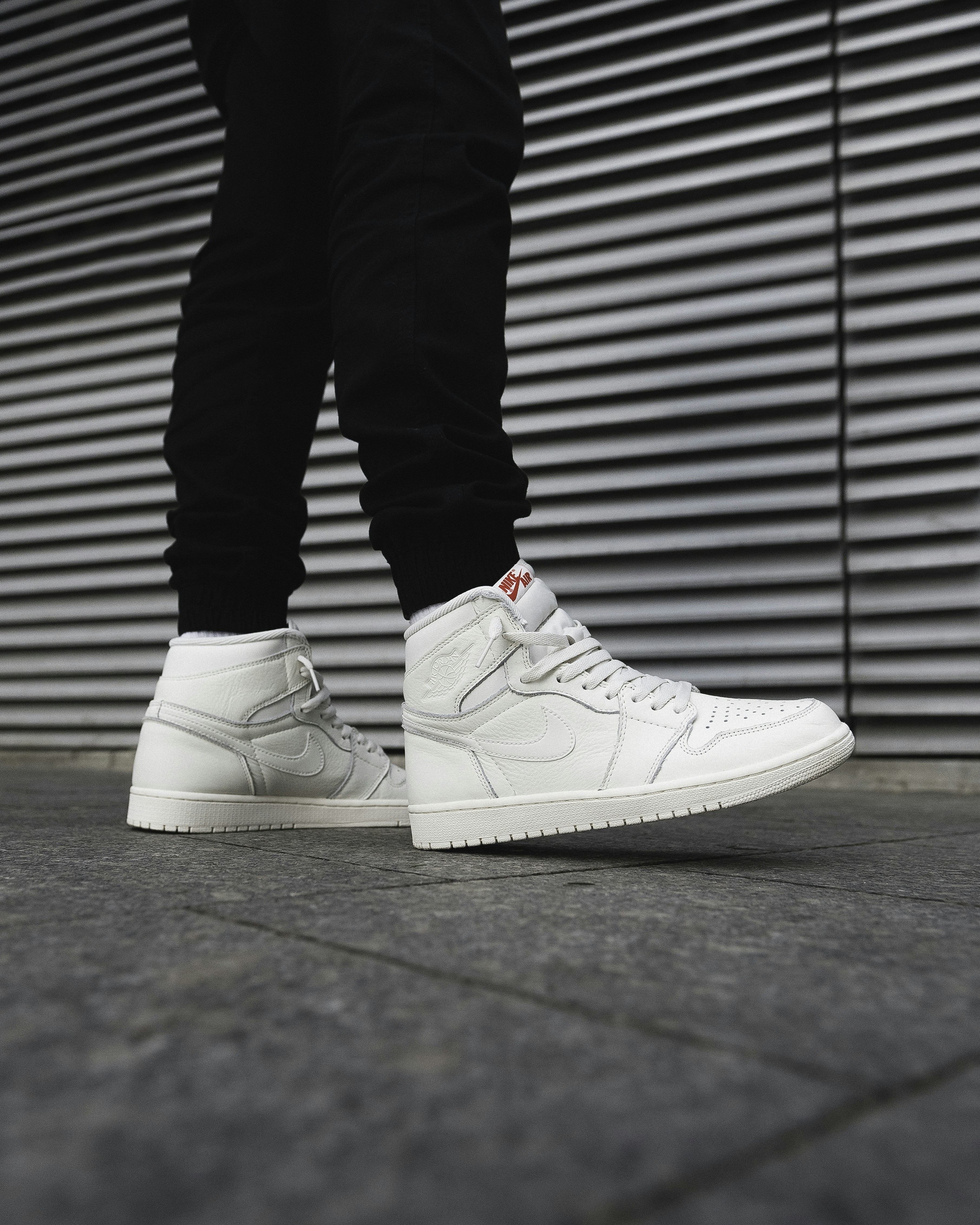 A person wearing white high-top sneakers and black trousers on a grey sidewalk