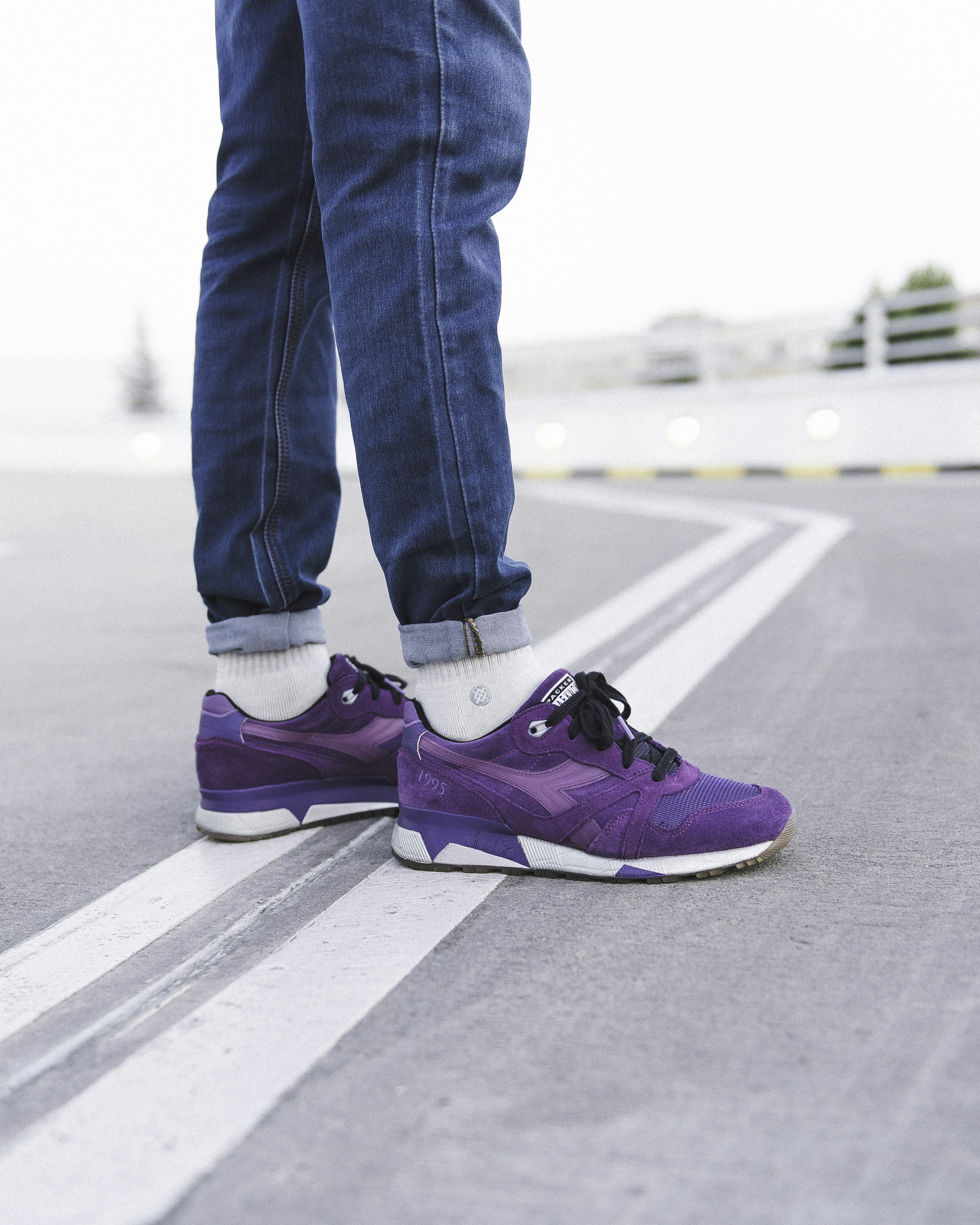 A person wearing blue jeans and purple suede sneakers on a paved road