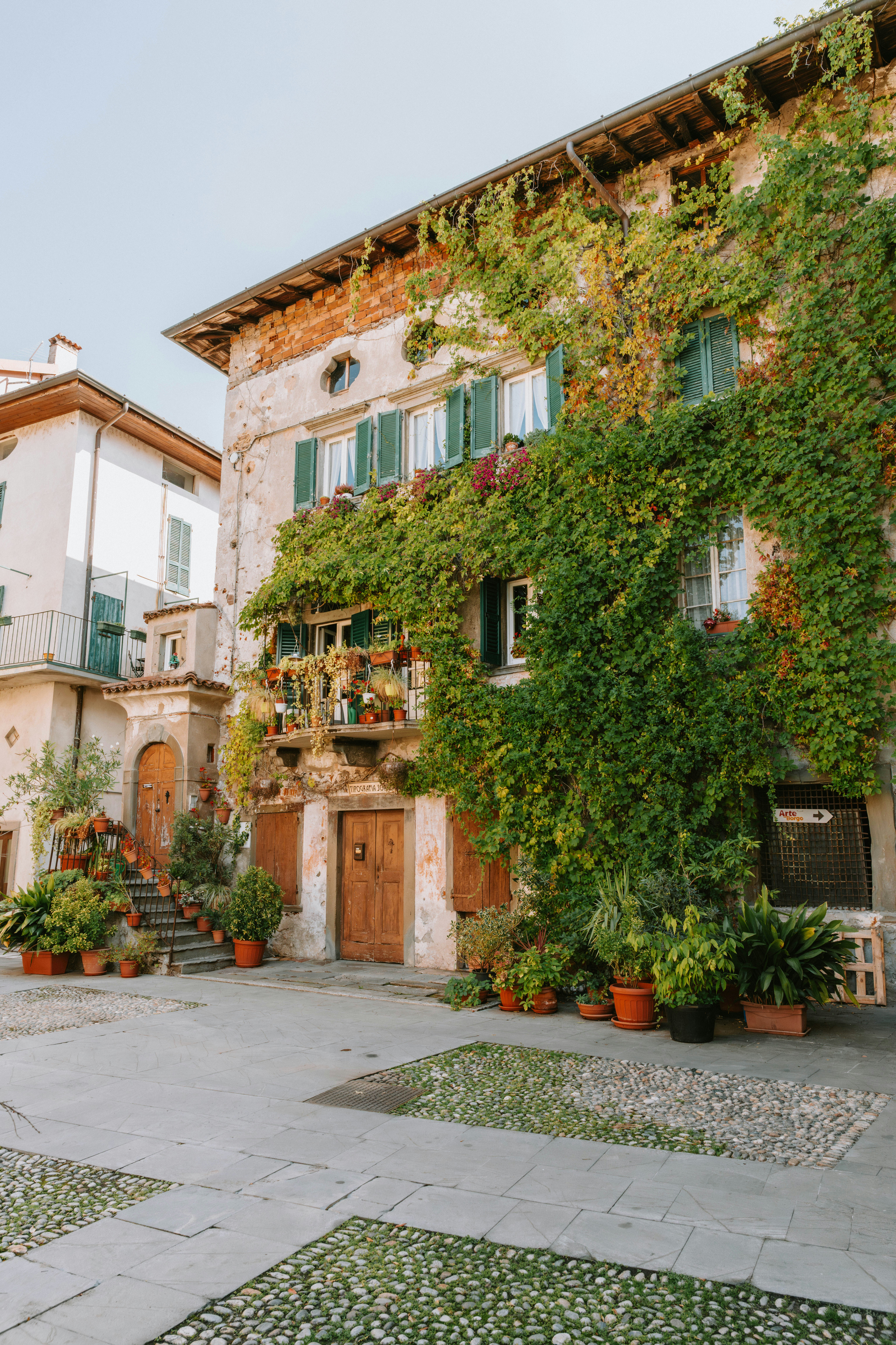 An old stone building covered in climbing vines with green shutters and potted plants