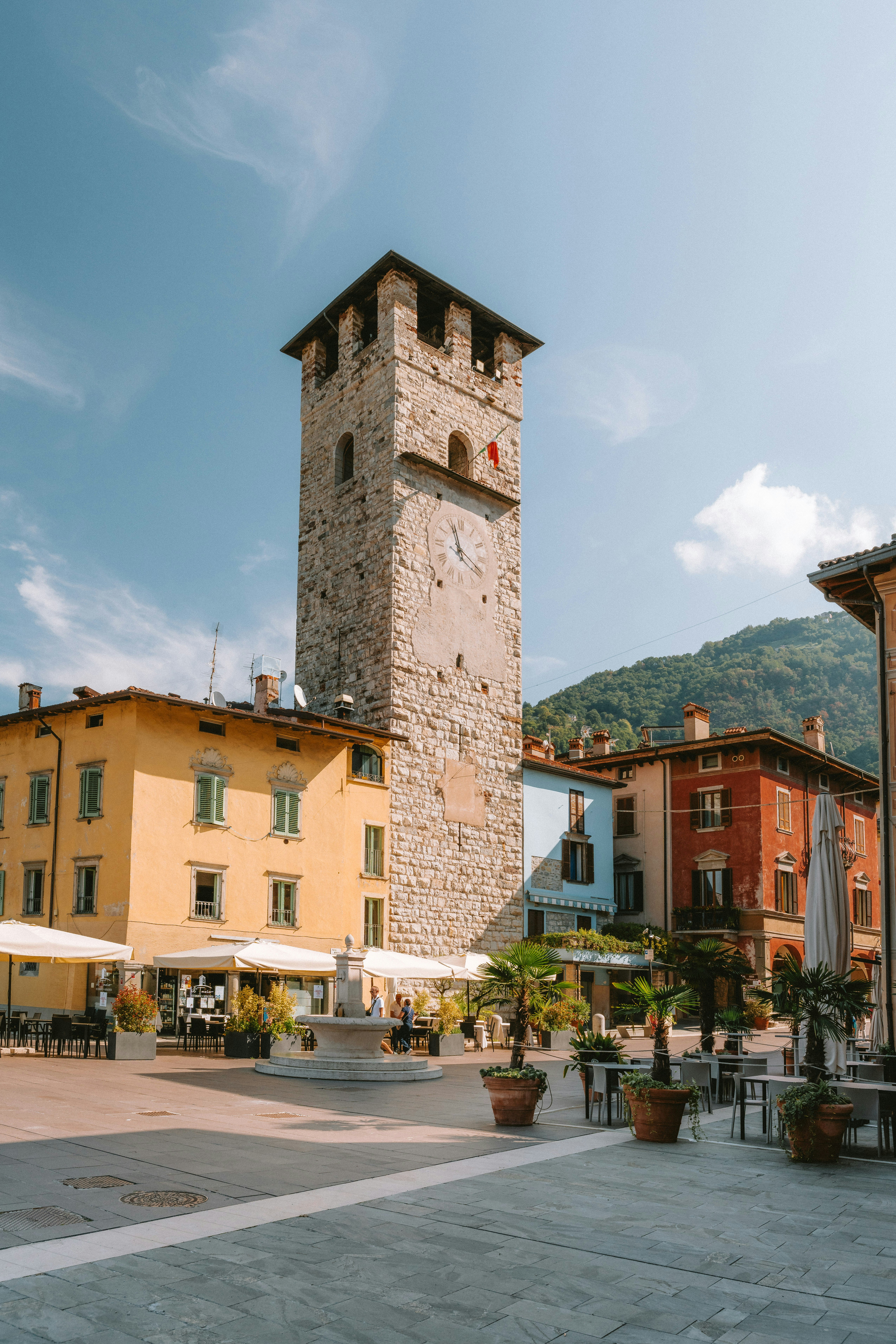 A stone clock tower in a town square with colorful buildings and palm trees