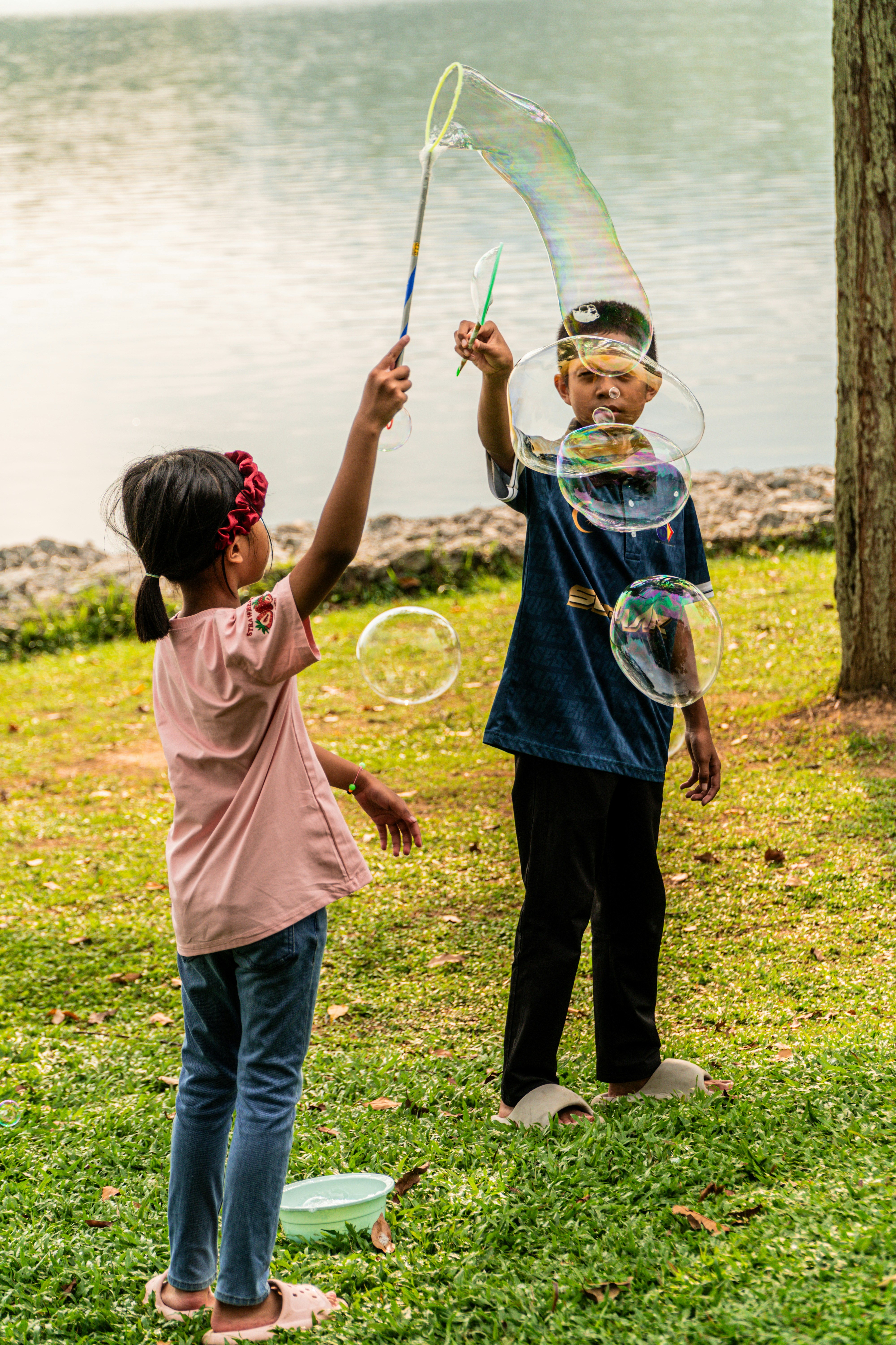 A girl and a boy making large bubbles on a grassy lakeshore
