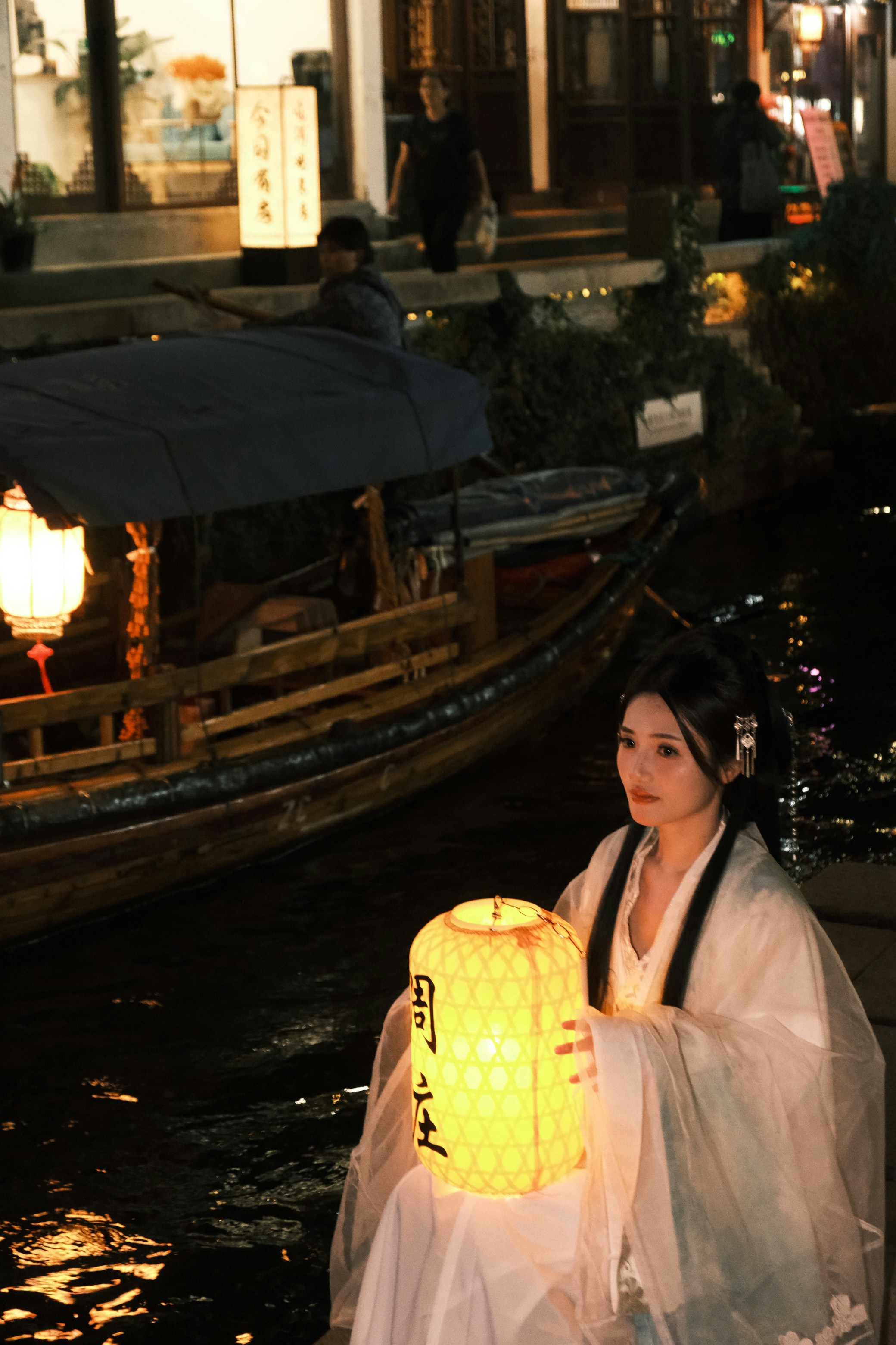 A woman in traditional white robes holding a glowing lantern beside a canal boat