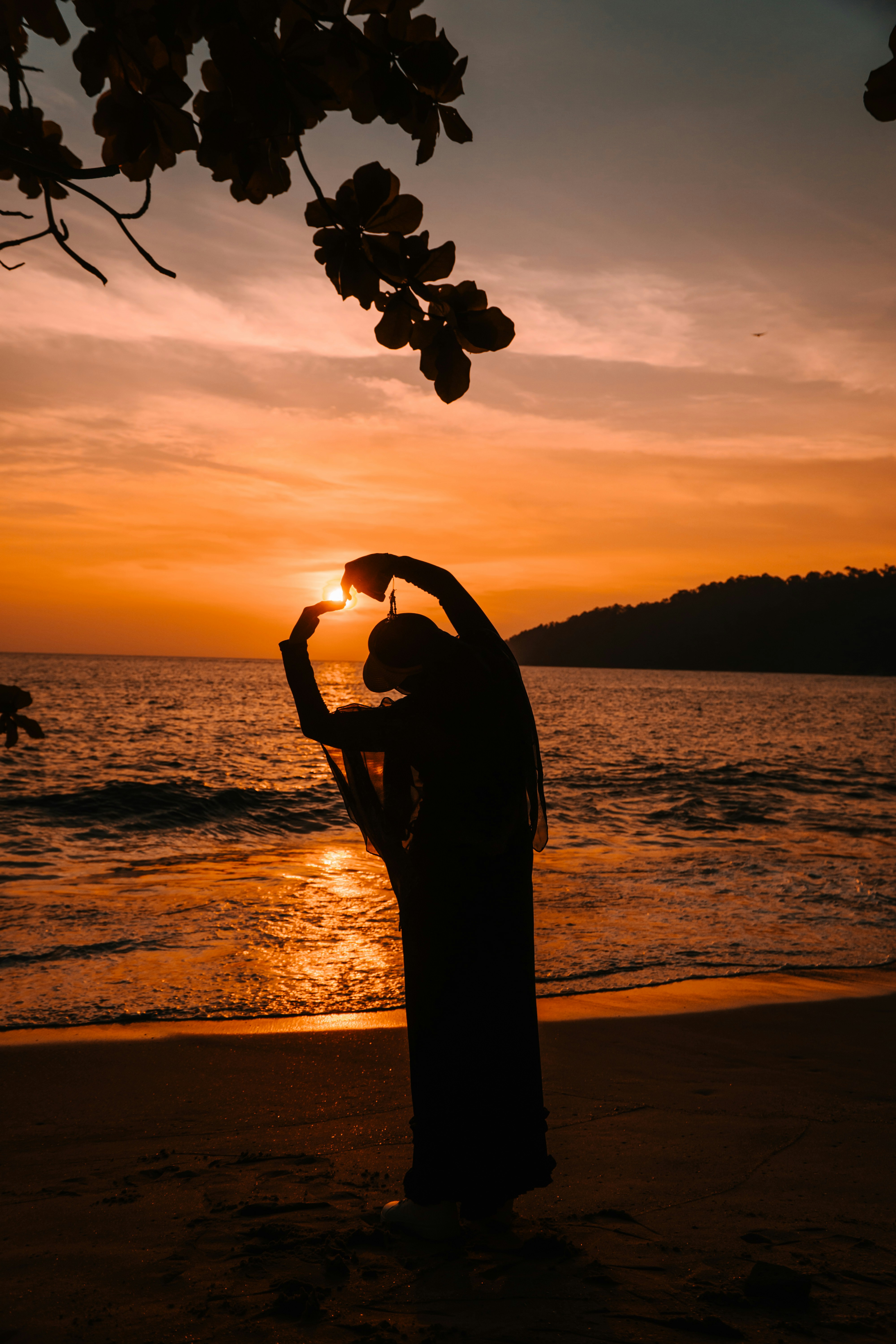 A person in silhouette framing the low sun with their hands on a beach