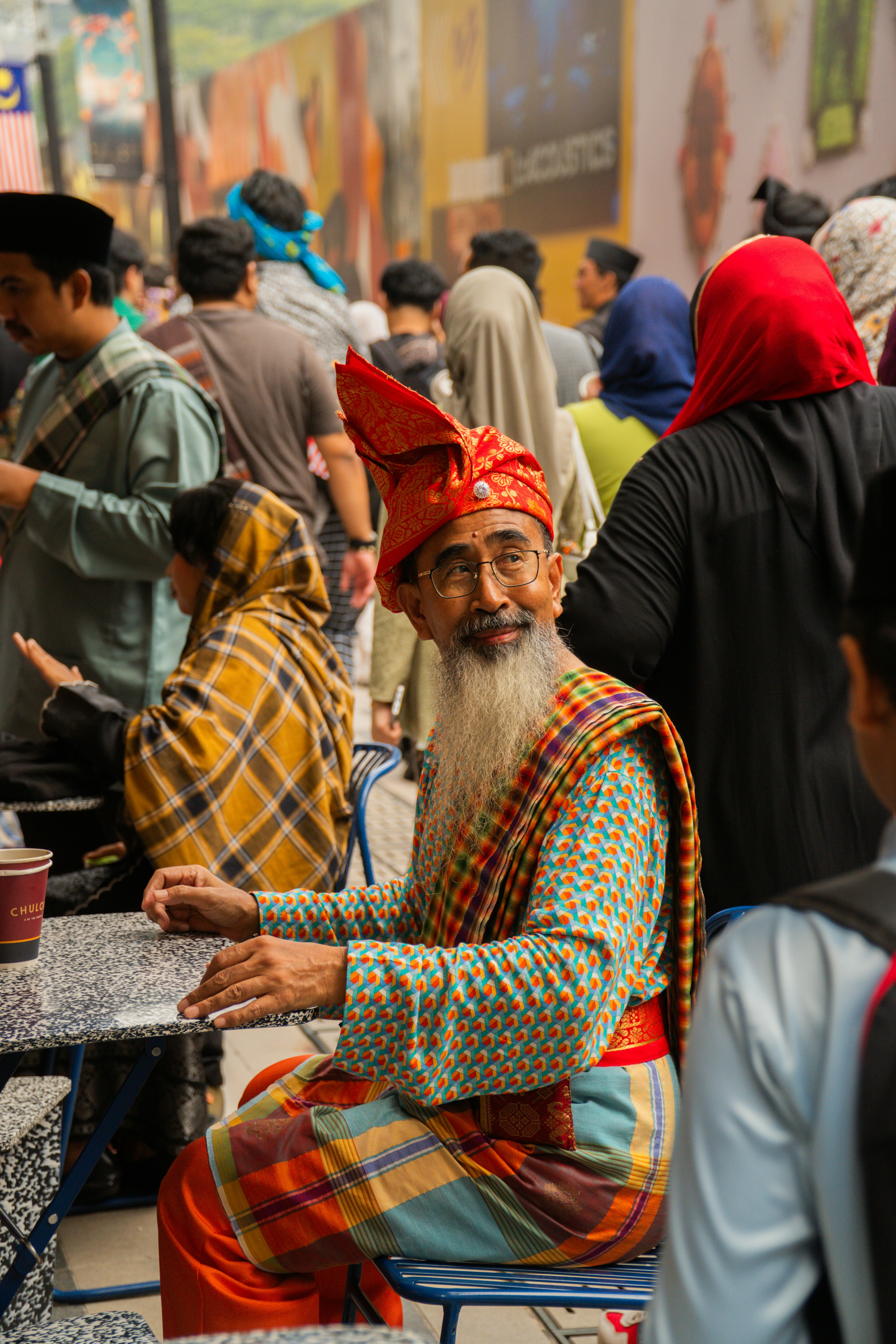 A bearded man in colorful traditional attire and red headdress among an outdoor crowd