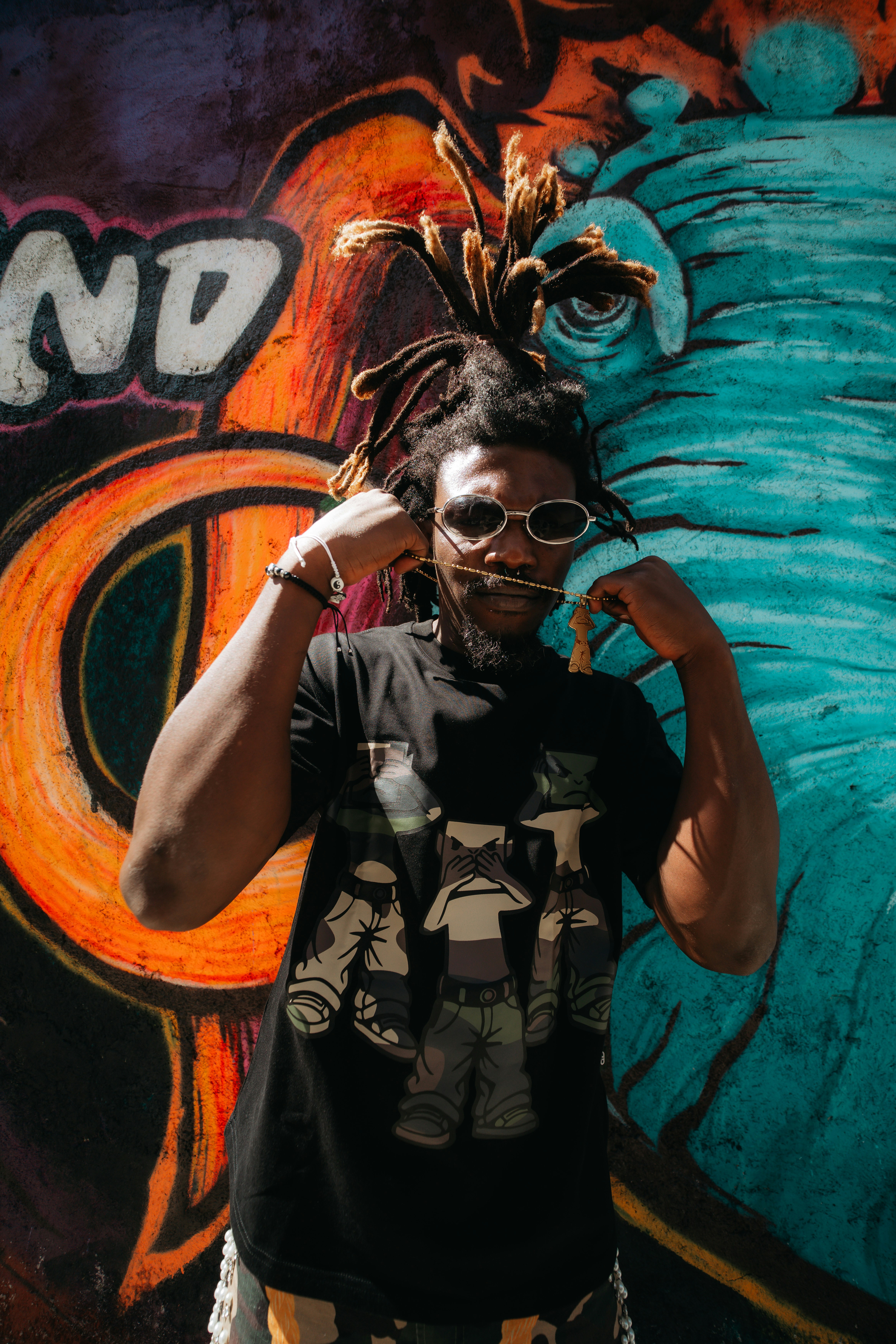 A person with dreadlocks wearing sunglasses and a graphic t-shirt against colorful graffiti