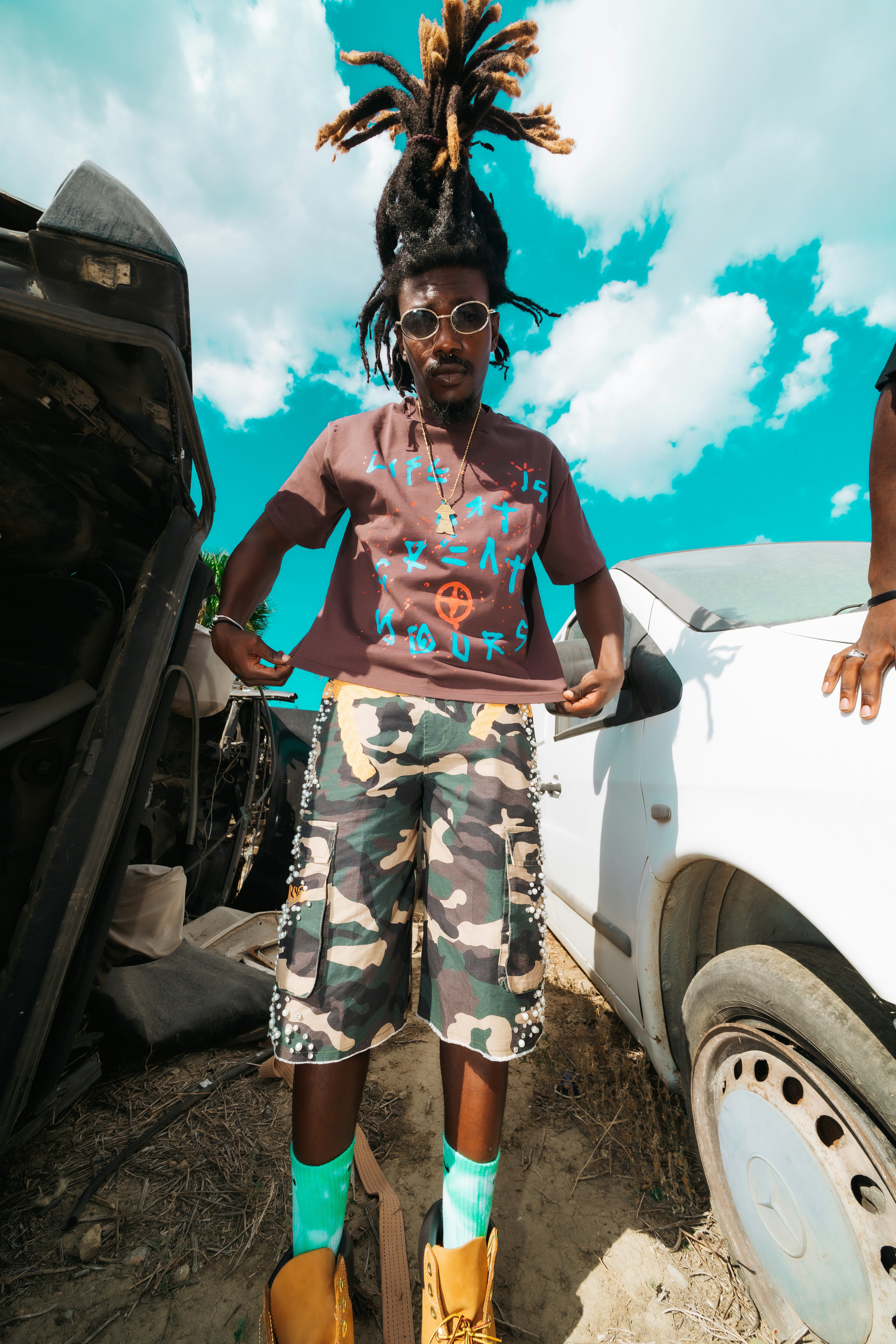 A person with dreadlocks wearing a brown t-shirt and camouflage shorts standing between two cars