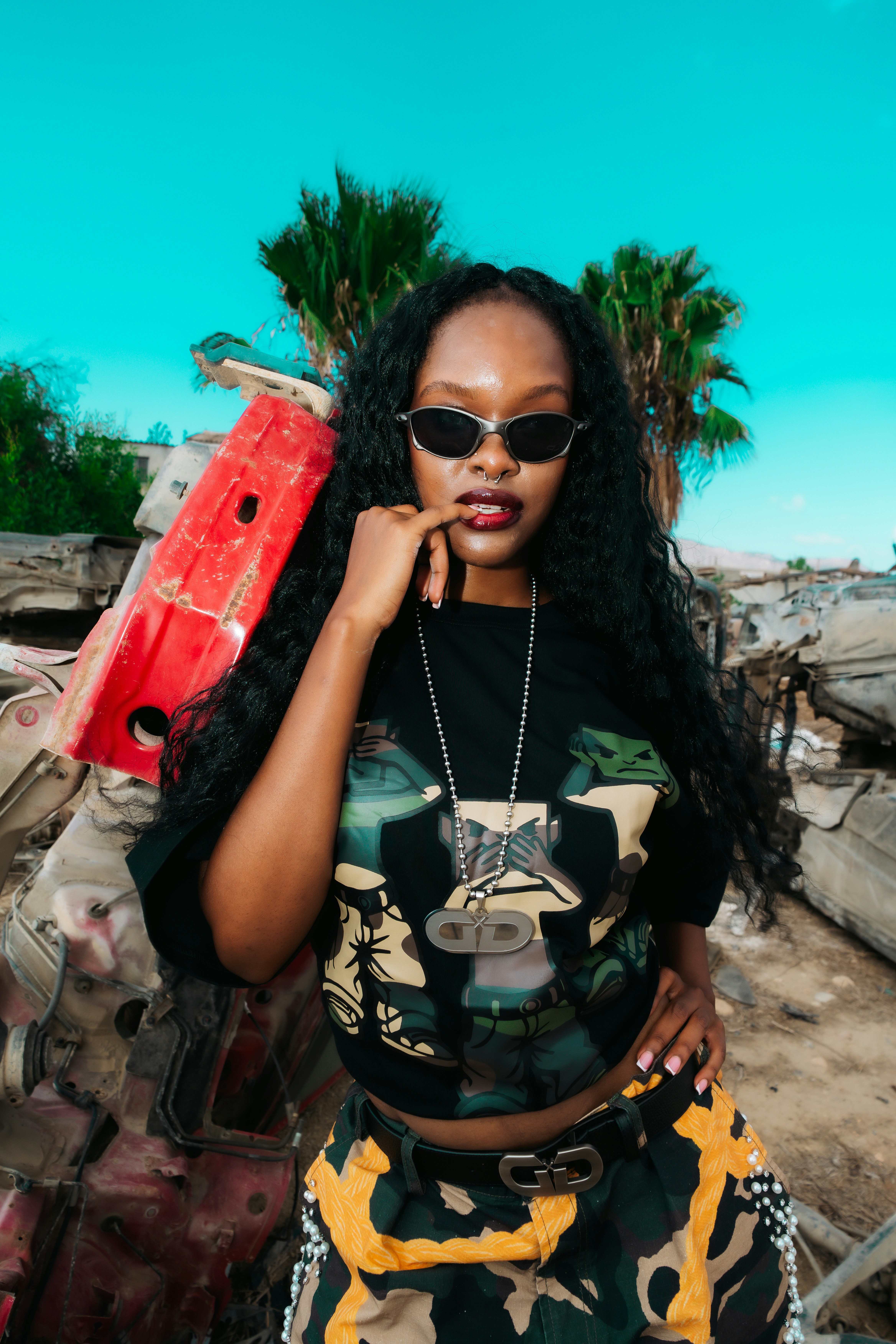 A woman with long dark hair wearing sunglasses and a graphic t-shirt in a junkyard