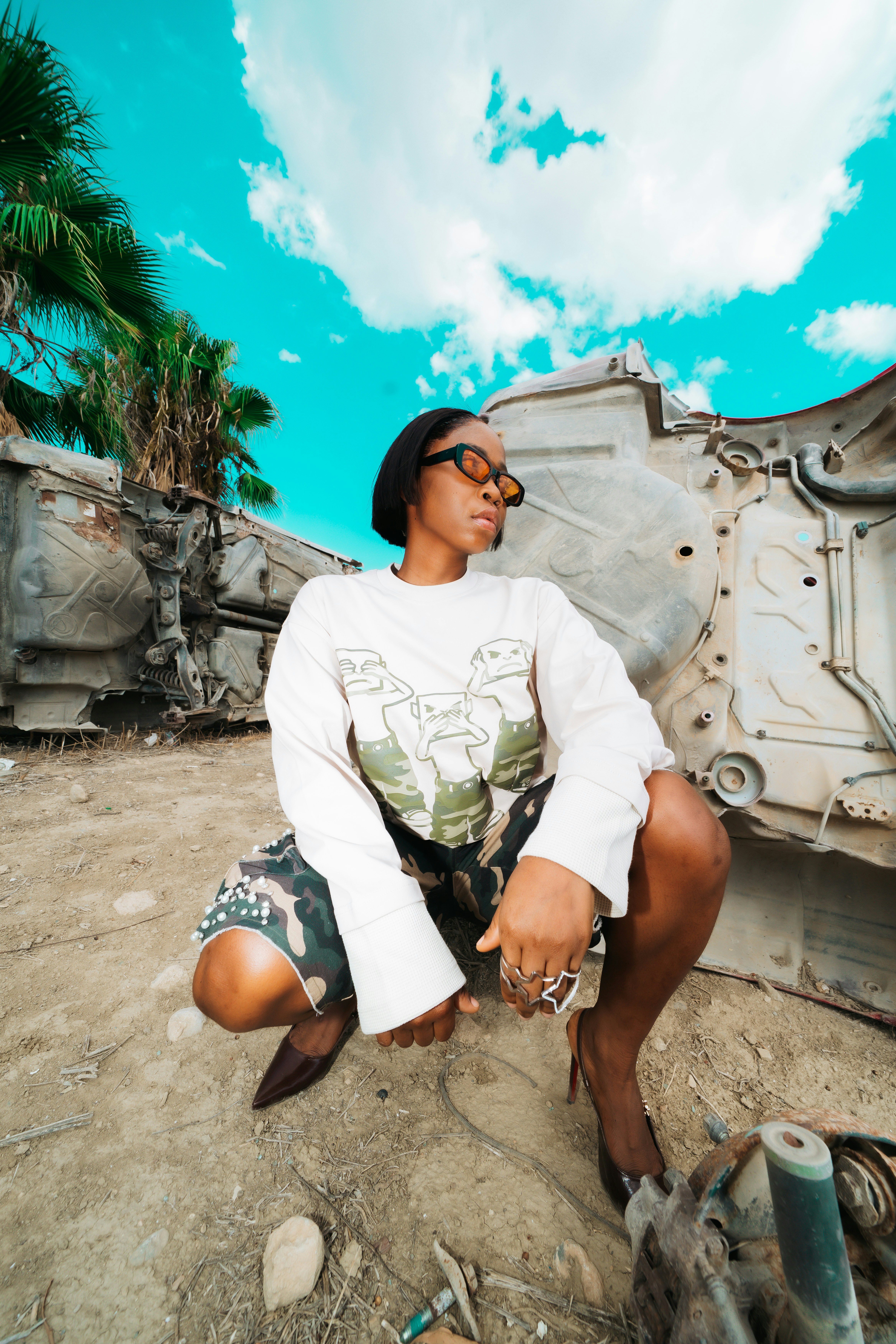 A person in a graphic sweatshirt and camouflage shorts crouching by a wrecked car