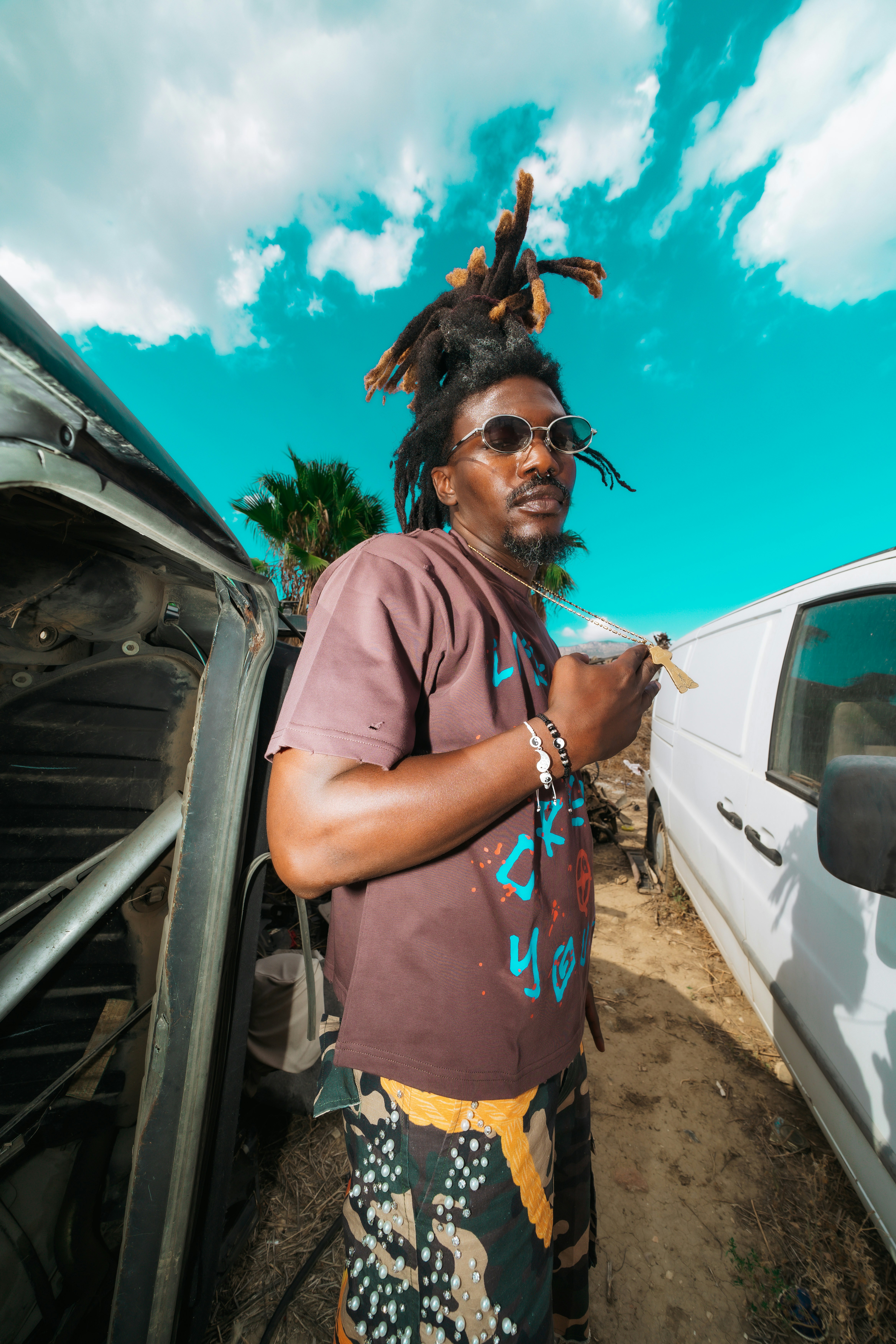A man with dreadlocks and sunglasses wearing a brown t-shirt between two vans