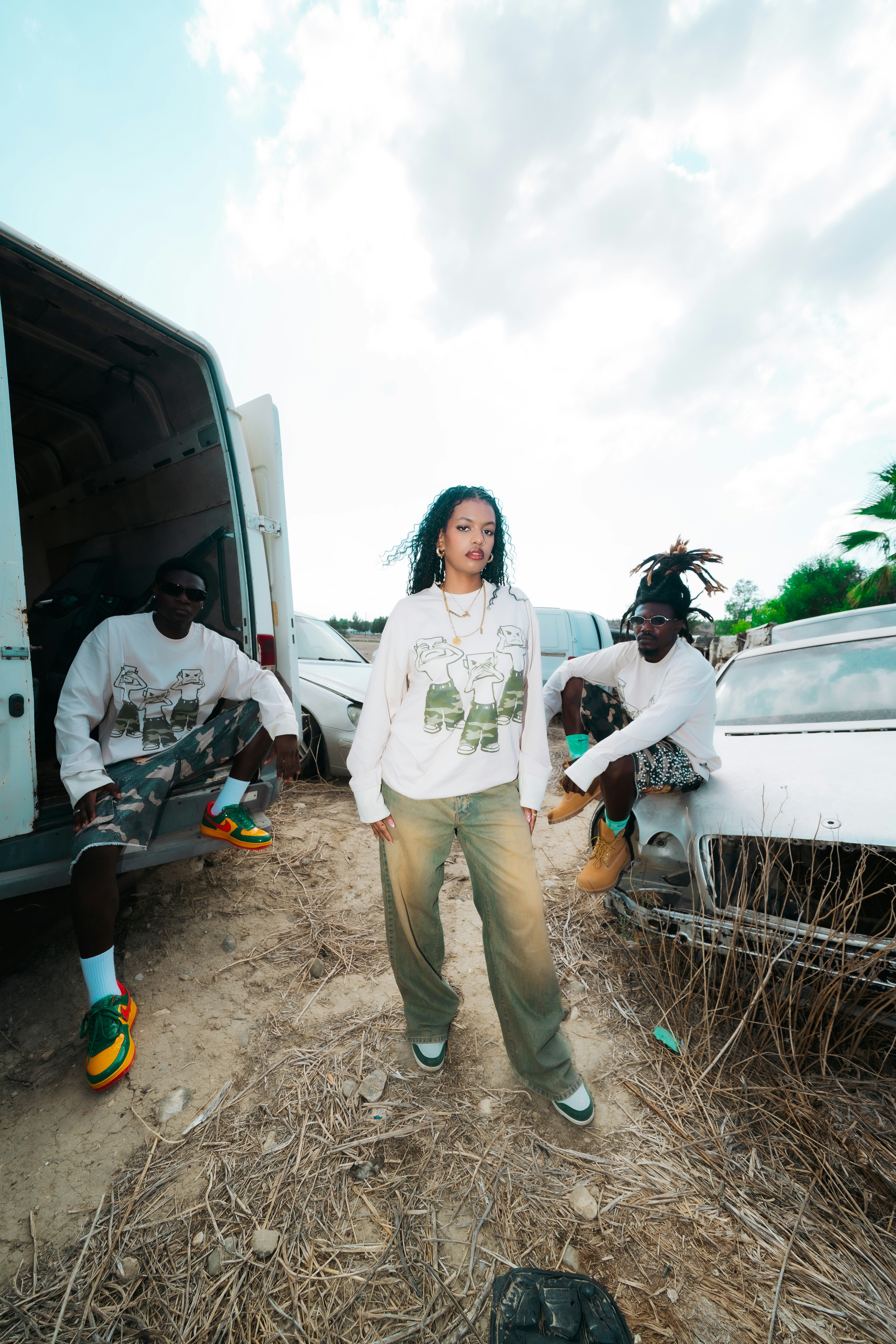 Three people in matching long-sleeved shirts standing among abandoned cars in a field