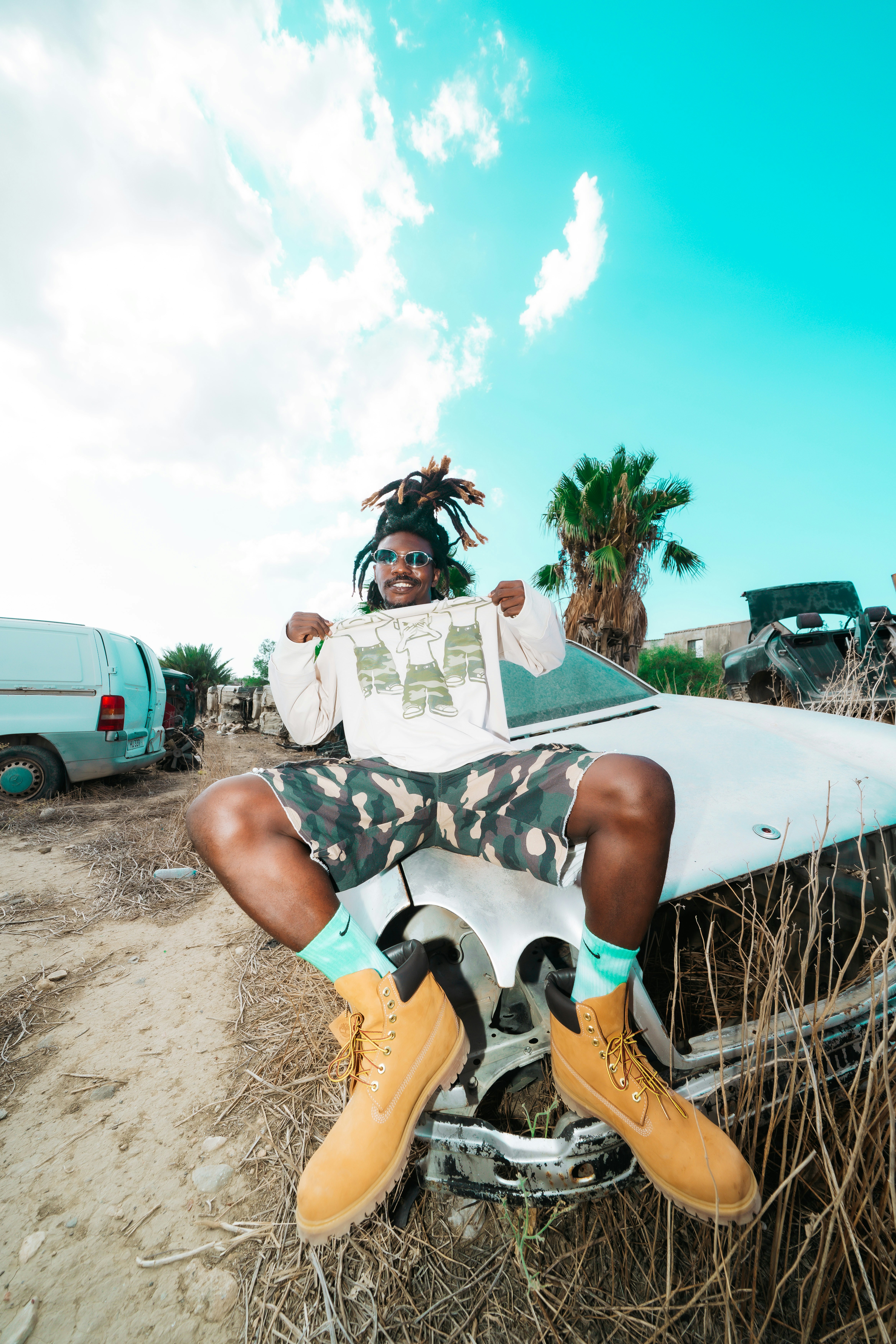 A person with dreadlocks wearing sunglasses and camouflage shorts sitting on a wrecked car