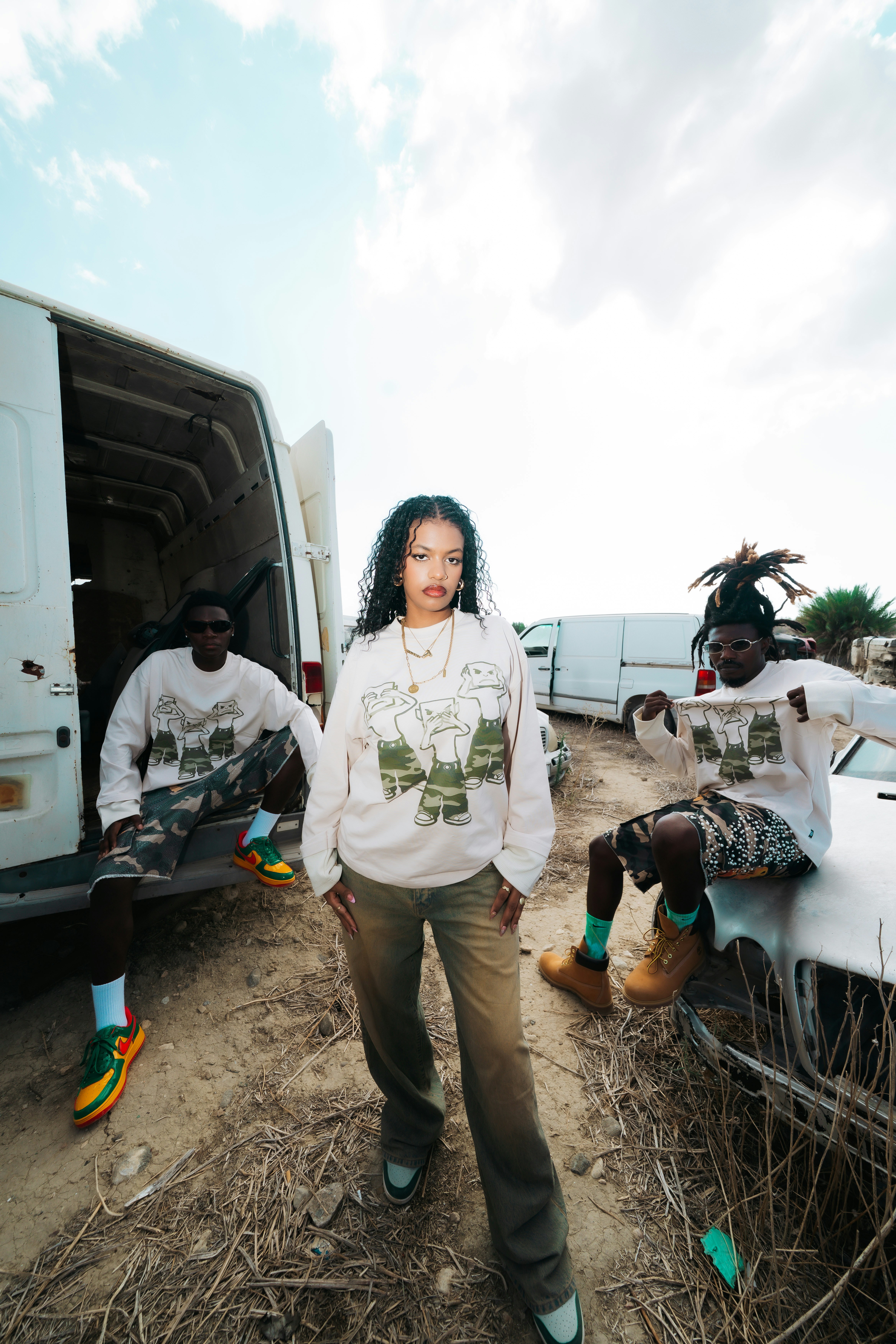 Three people in matching graphic sweatshirts standing near old vehicles in a dry field