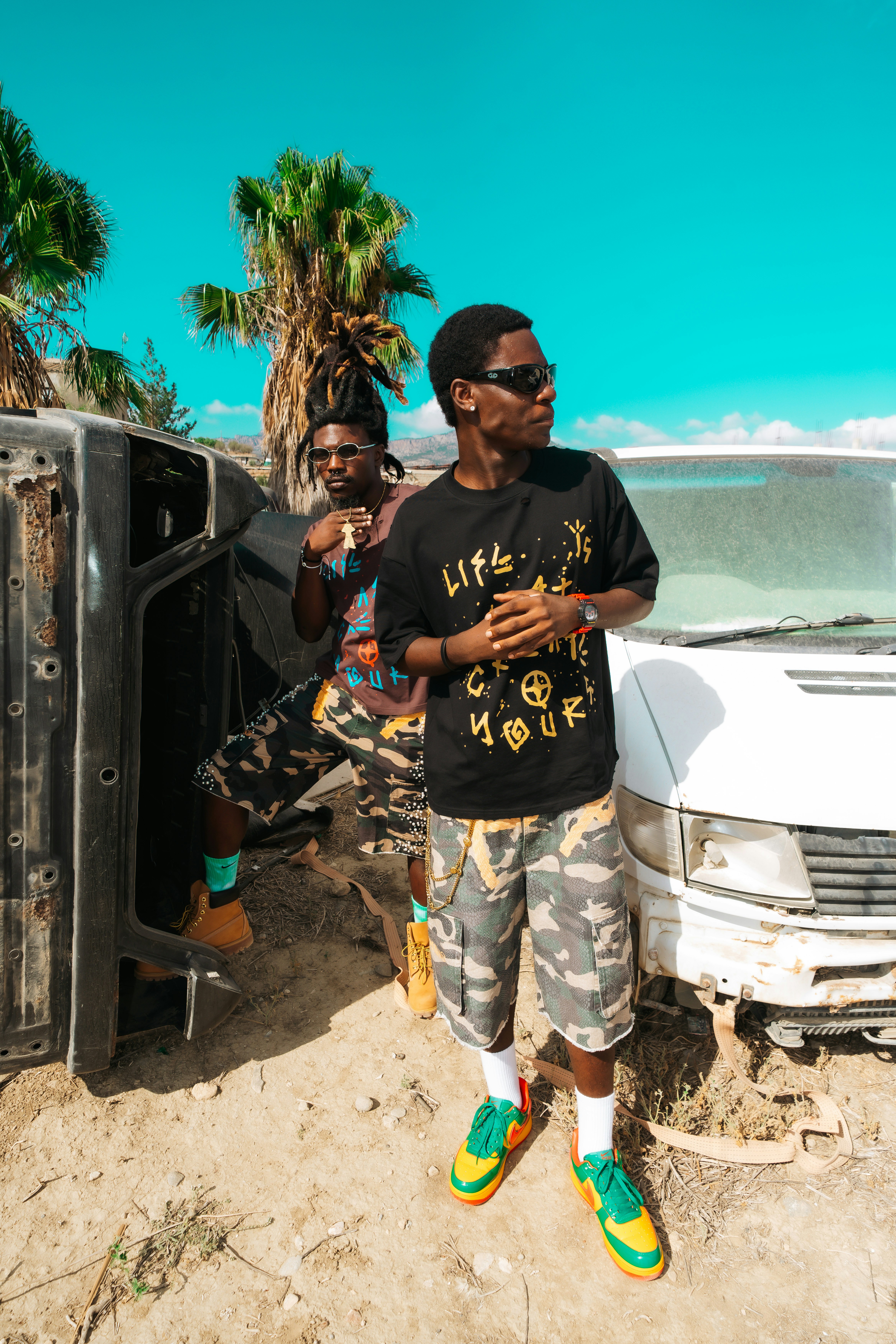 Two men in camouflage shorts and graphic t-shirts standing near a wrecked white van