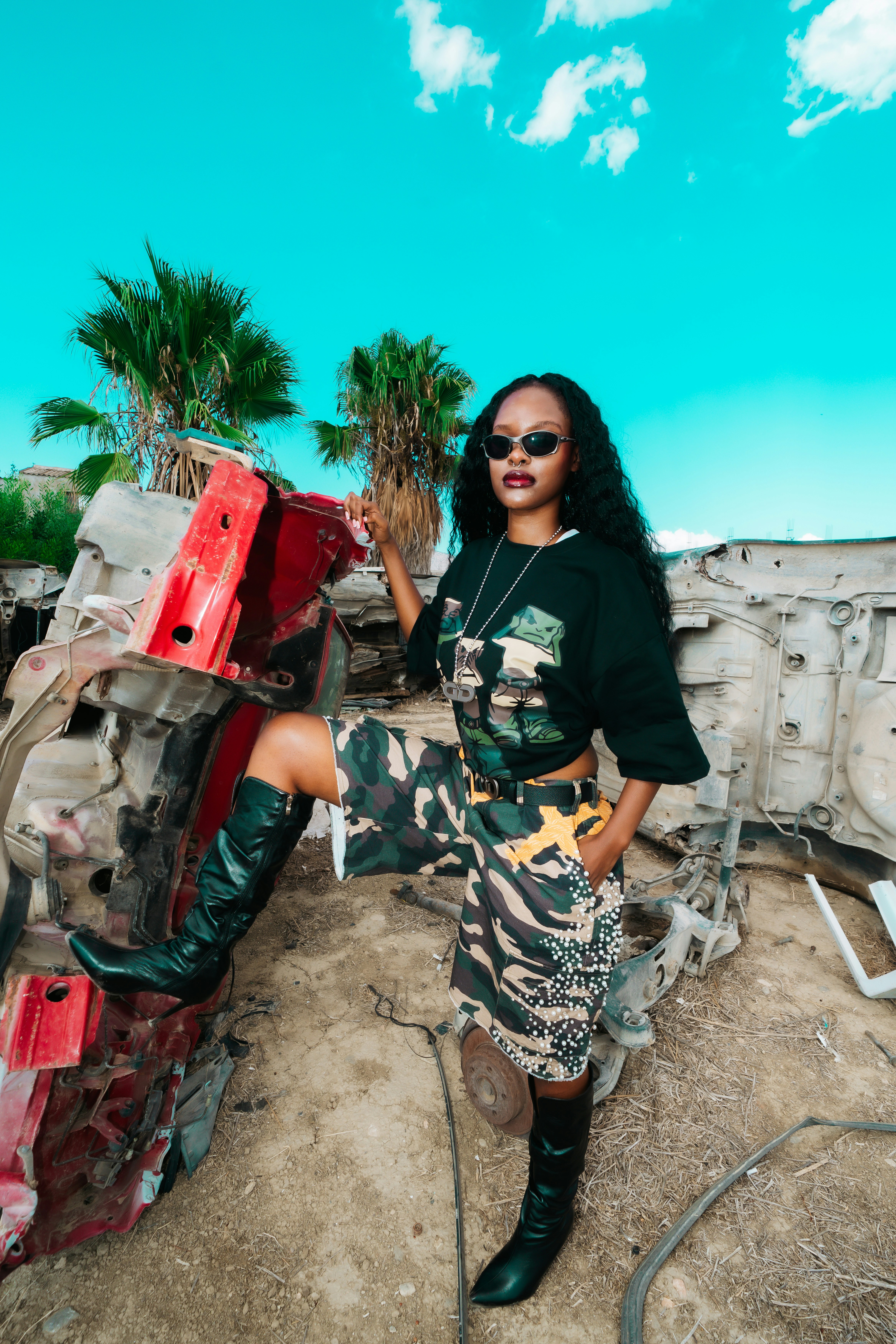 A woman in a graphic t-shirt and camouflage shorts posing by a wrecked car