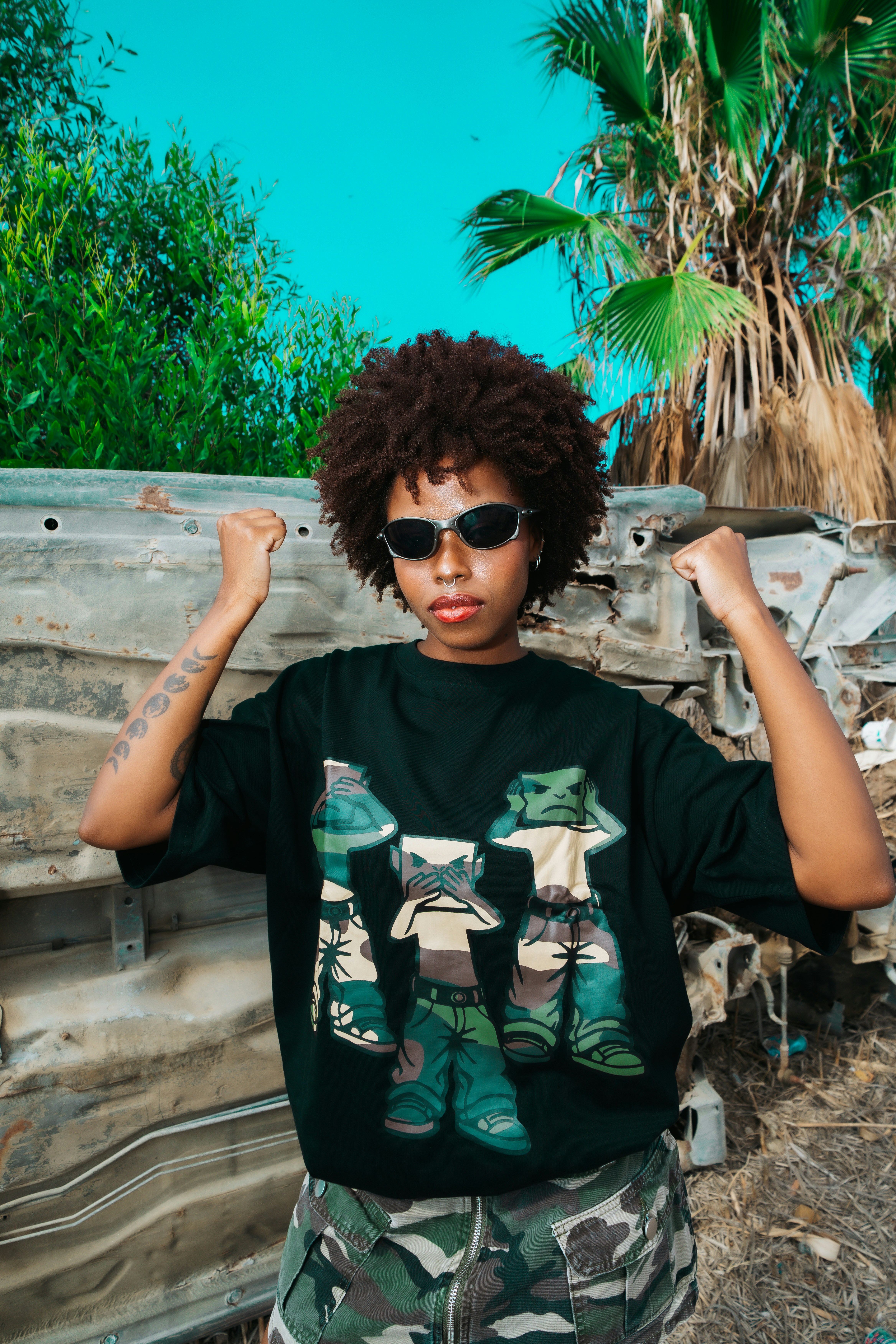 A person with curly hair and sunglasses flexing arms in front of a car frame