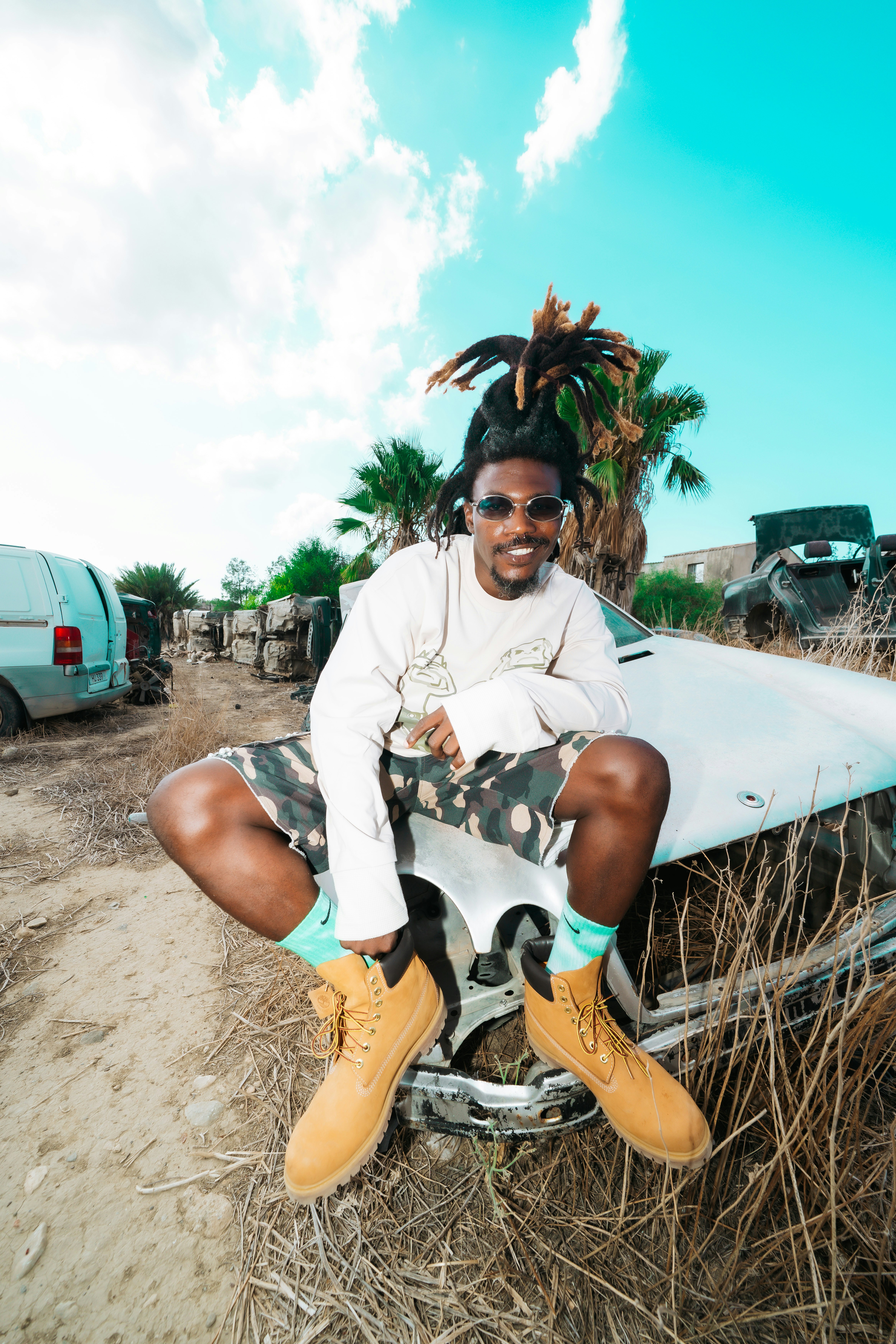 A person with dreadlocks wearing sunglasses and camouflage shorts sitting on a wrecked car