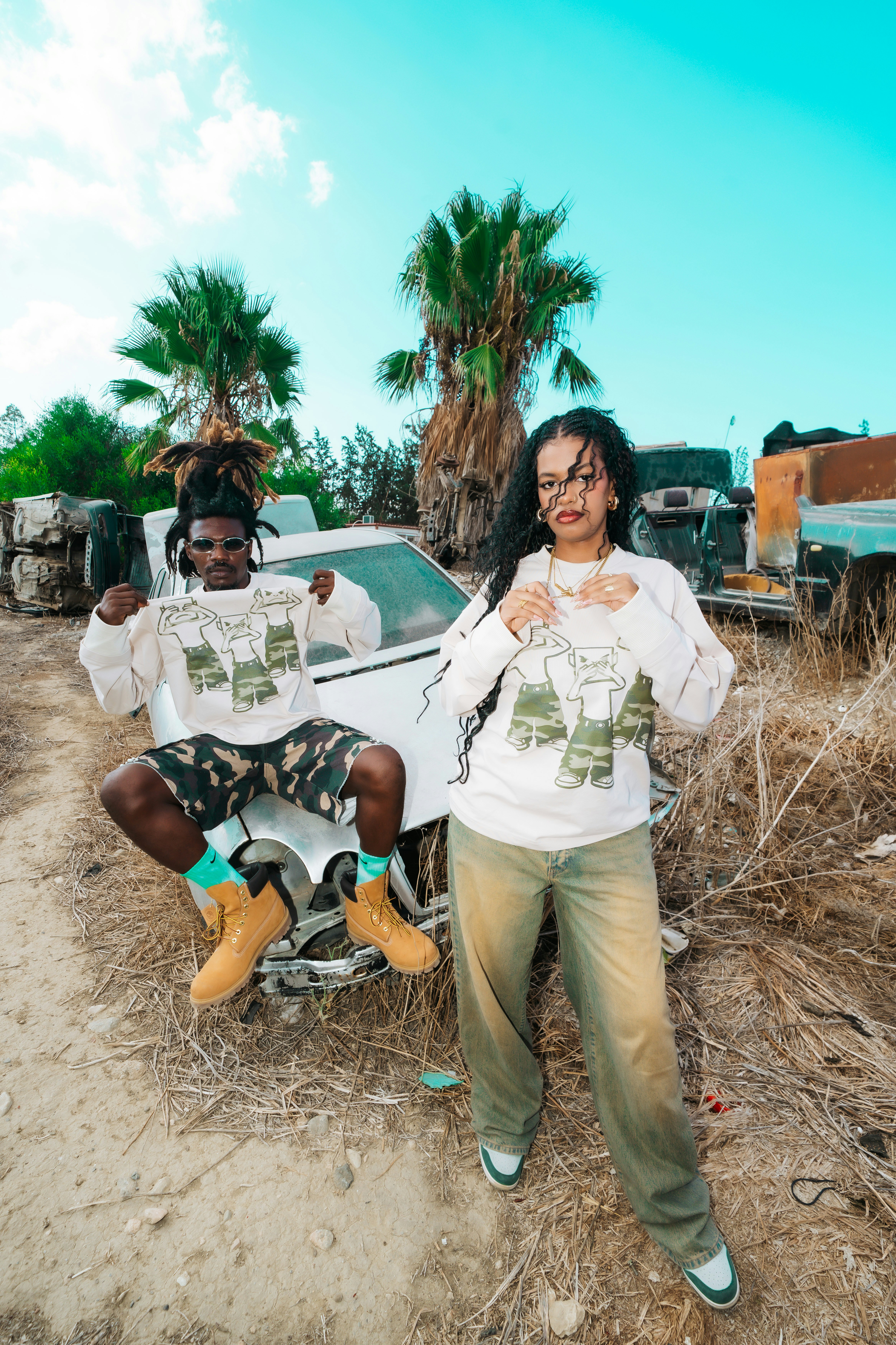 A man and a woman in matching graphic sweatshirts standing by a wrecked car