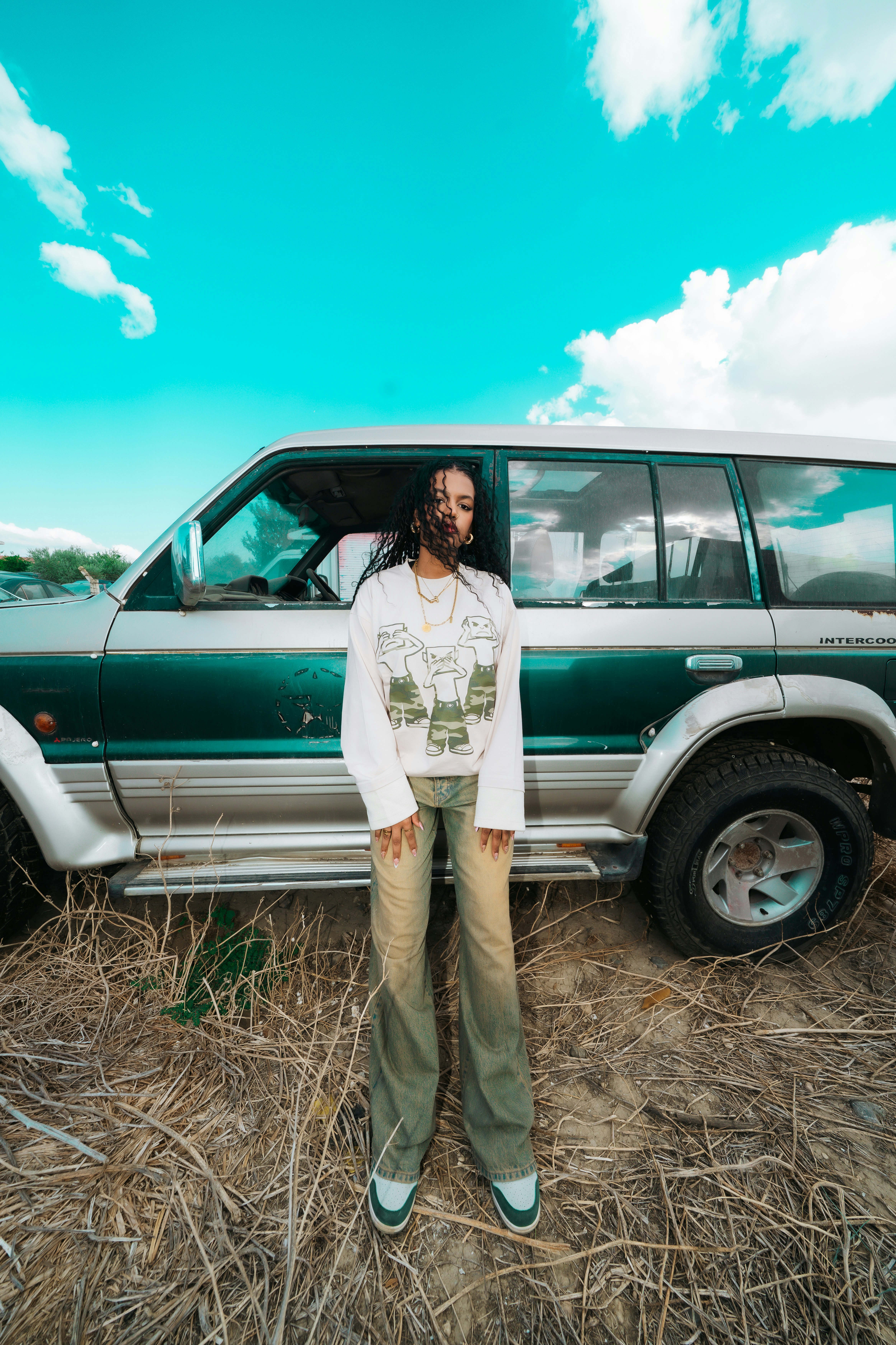 A person in a graphic sweatshirt and jeans standing before a green and silver SUV