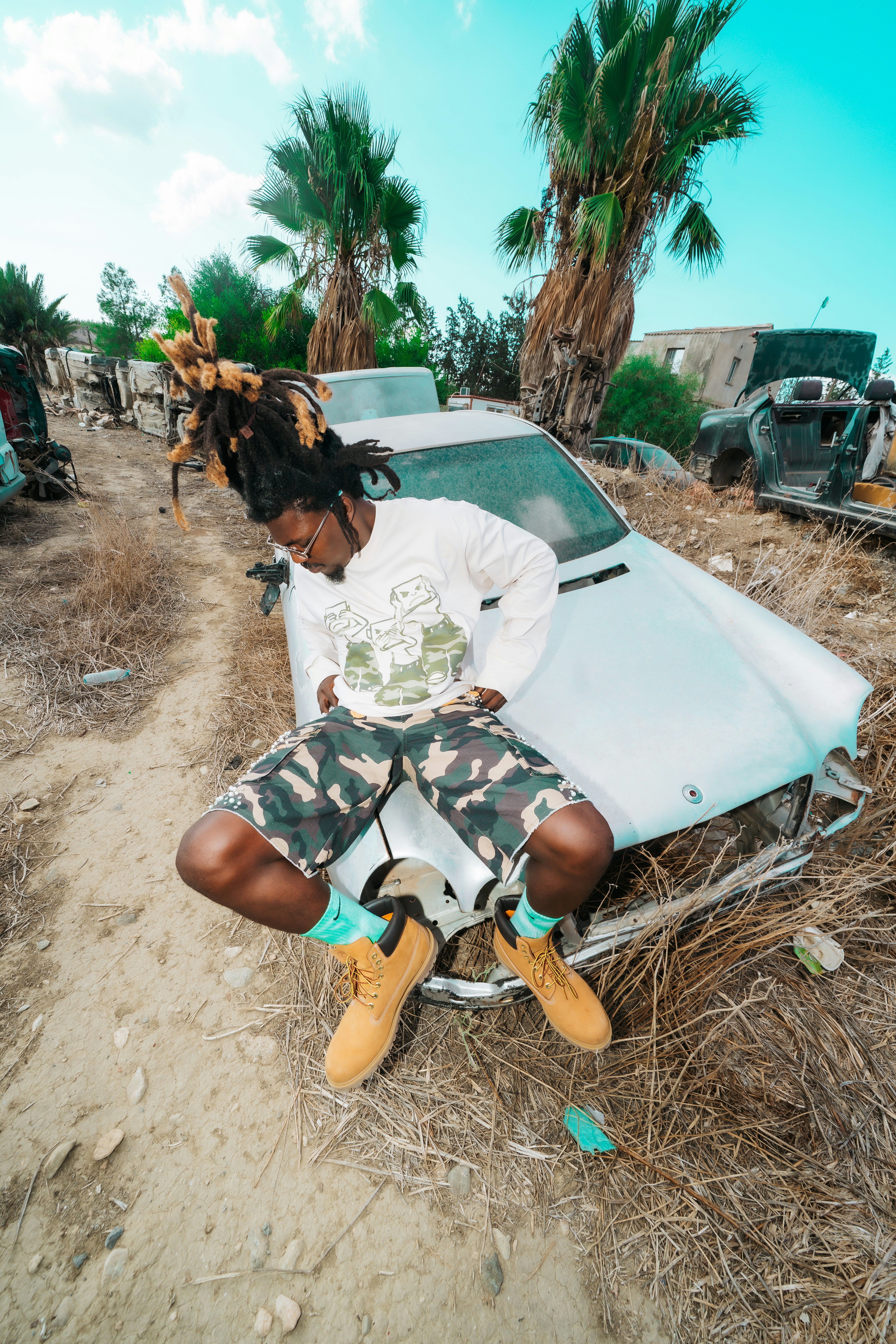 A person with dreadlocks sitting on a wrecked car in a field with palm trees