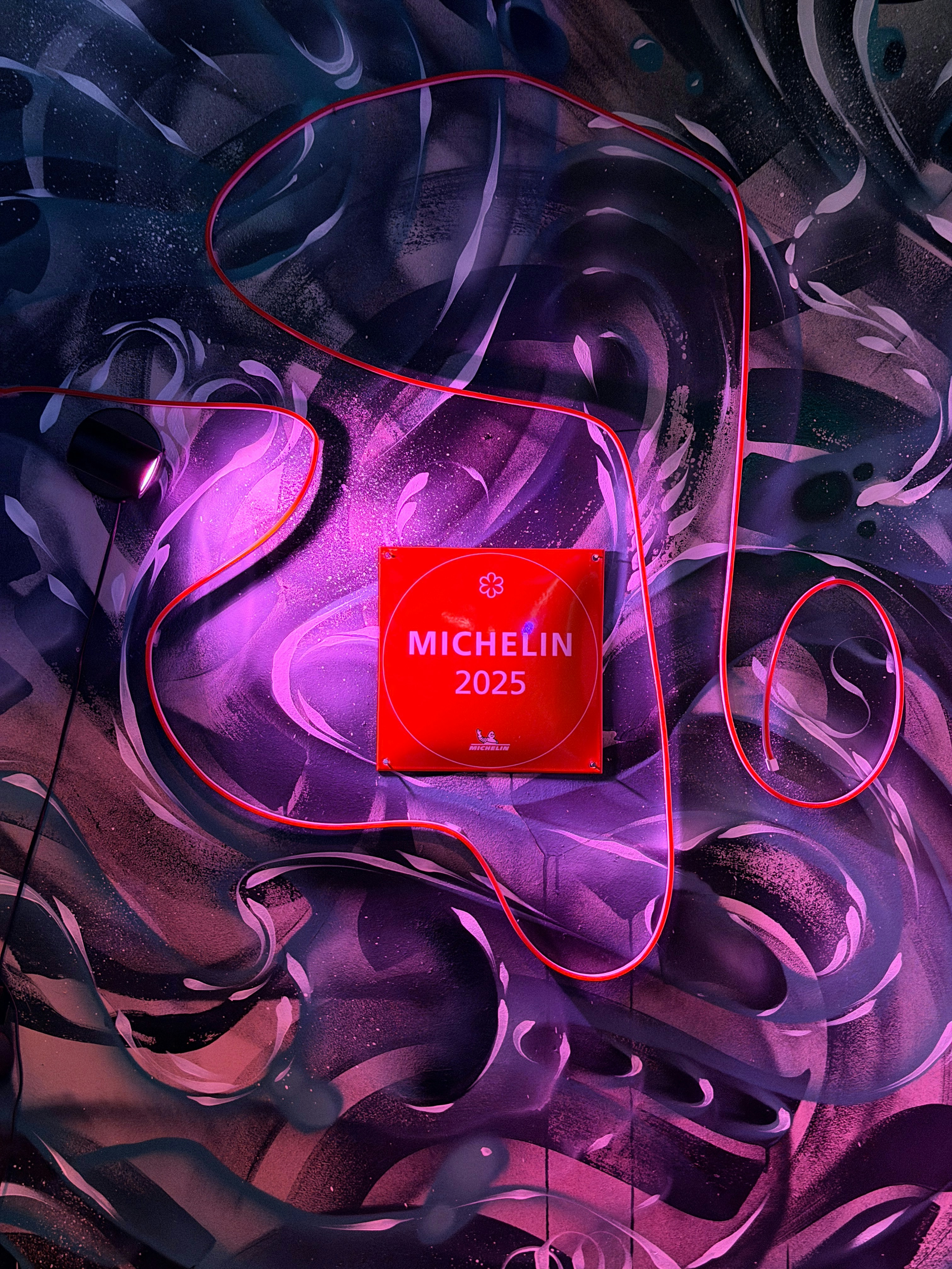 A red Michelin 2025 plaque surrounded by a glowing red neon tube on a mural