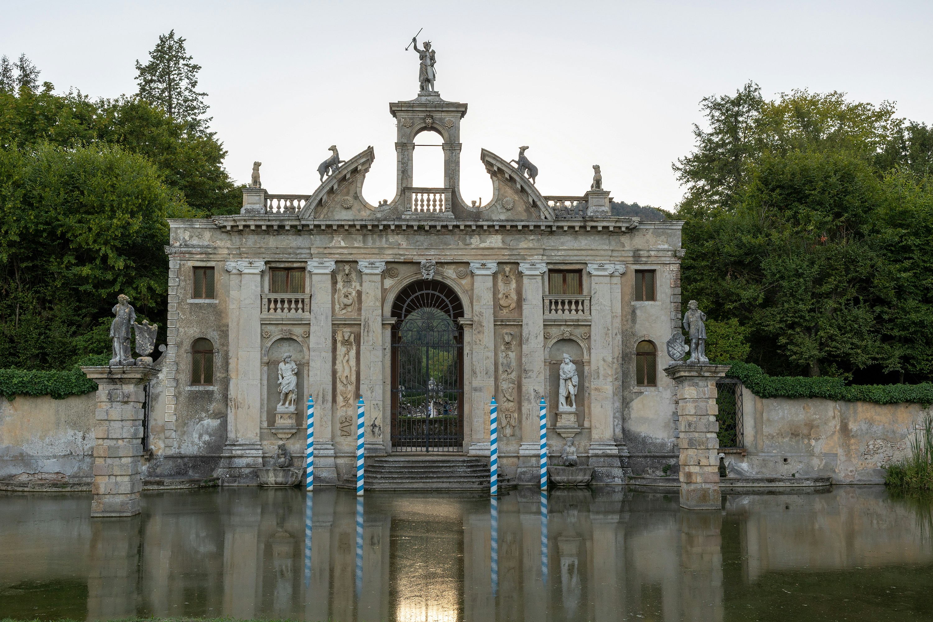An ornate stone villa entrance with statues and blue striped poles by a pond