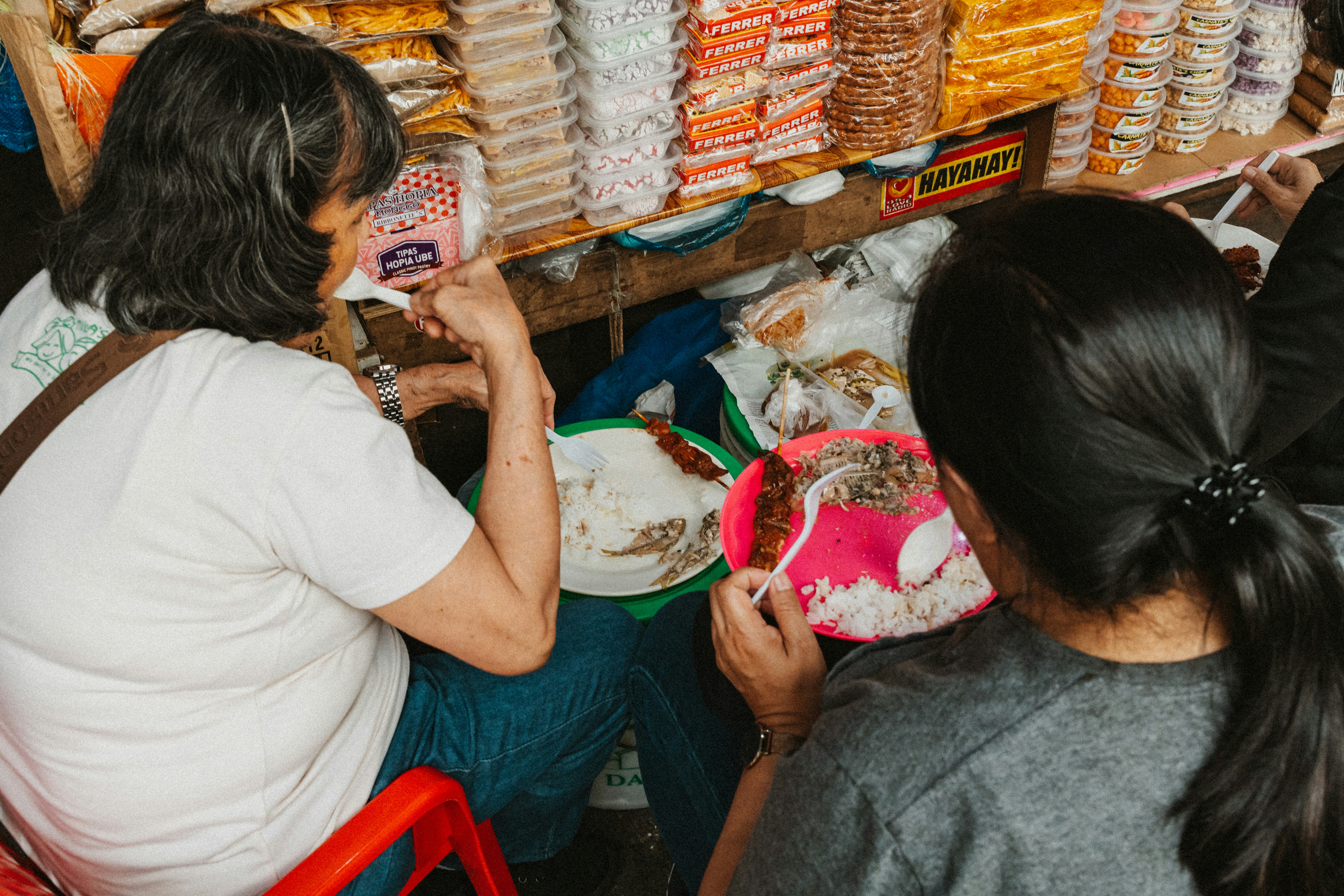 Two people eating rice and meat skewers at a market stall with snacks