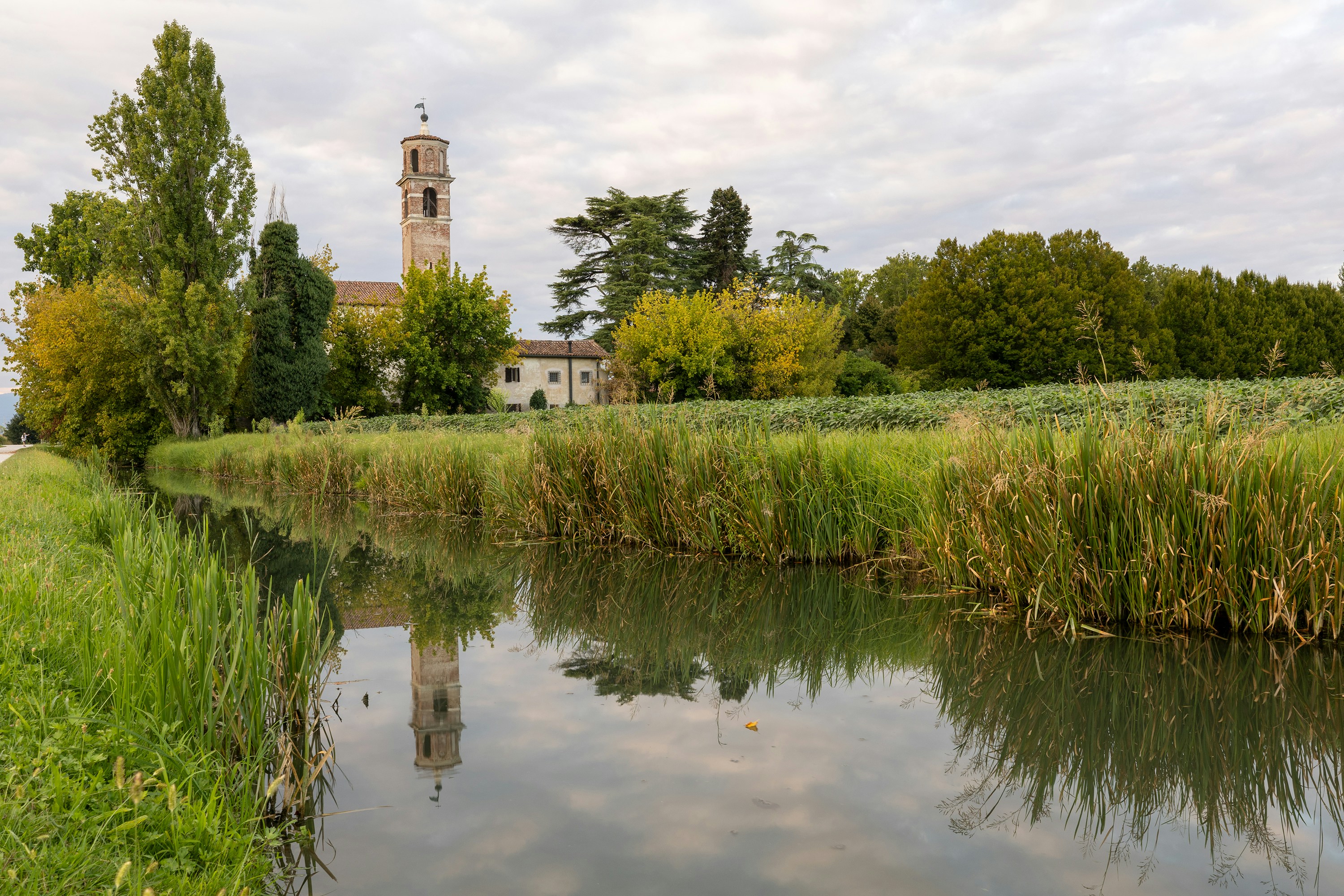 A stone bell tower reflecting in a calm canal beside green trees and fields