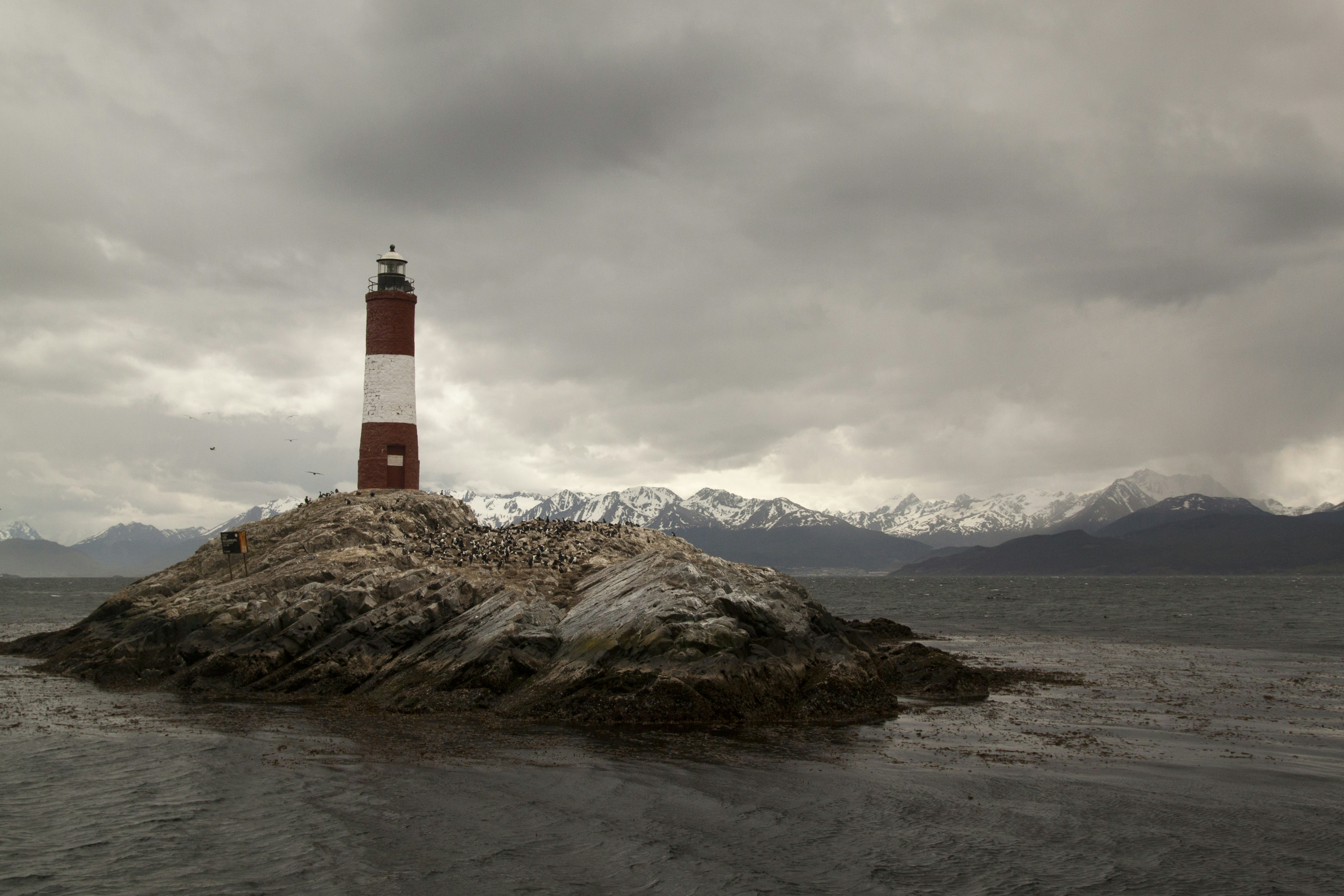 A red and white lighthouse on a rocky island with snow-capped mountains behind