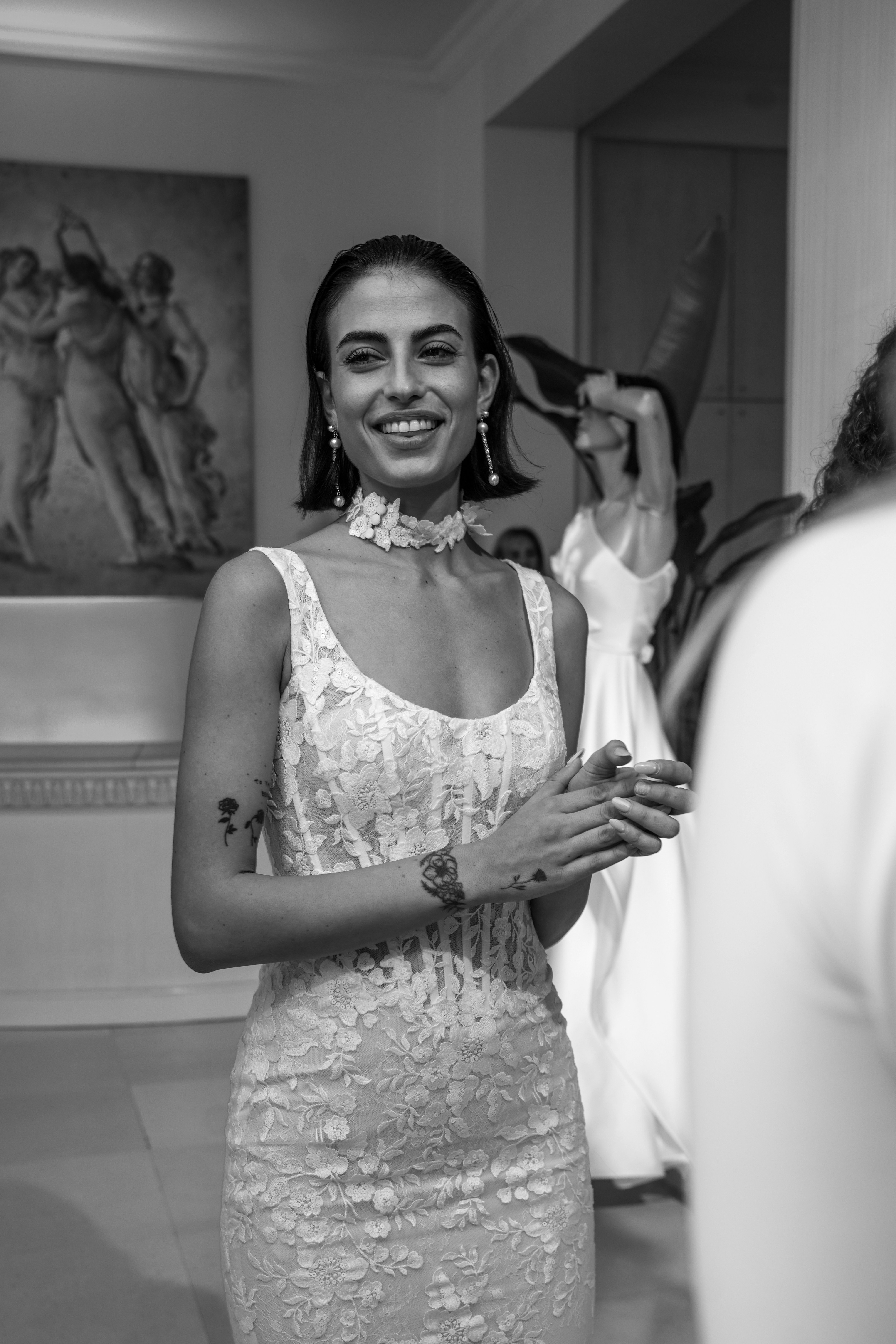 A smiling woman in a white lace dress and floral choker in a gallery