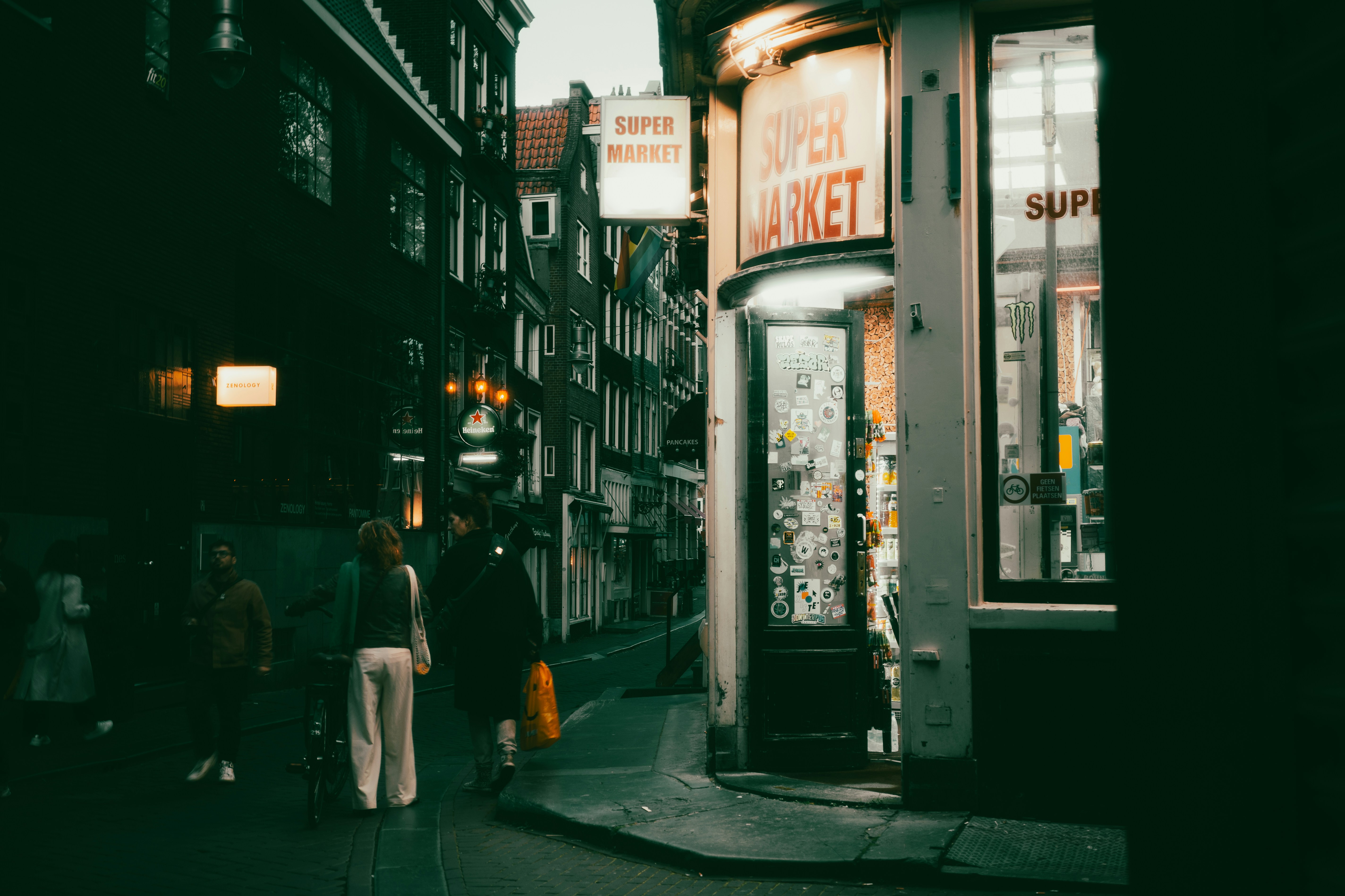 People walking past a corner supermarket with a sticker-covered door on a city street
