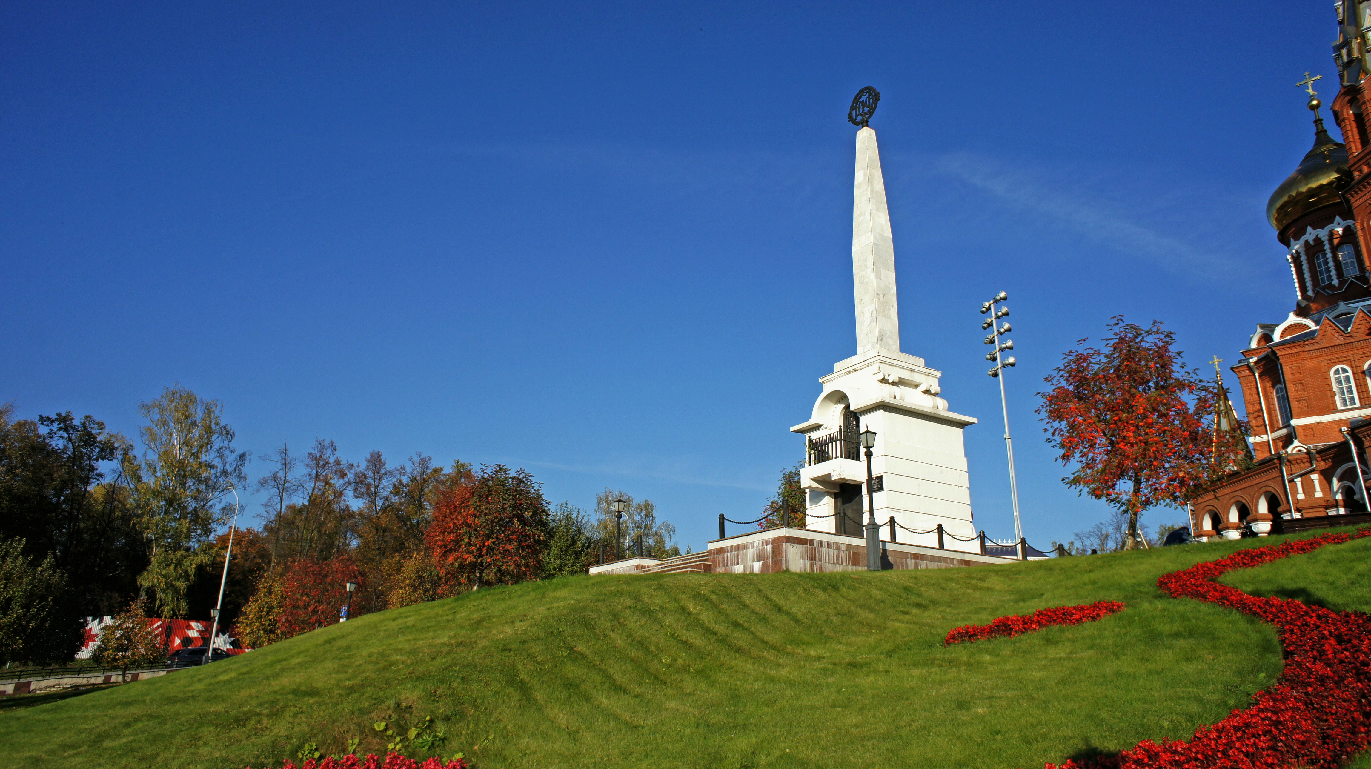 A white stone obelisk monument on a grassy hill beside a red brick church
